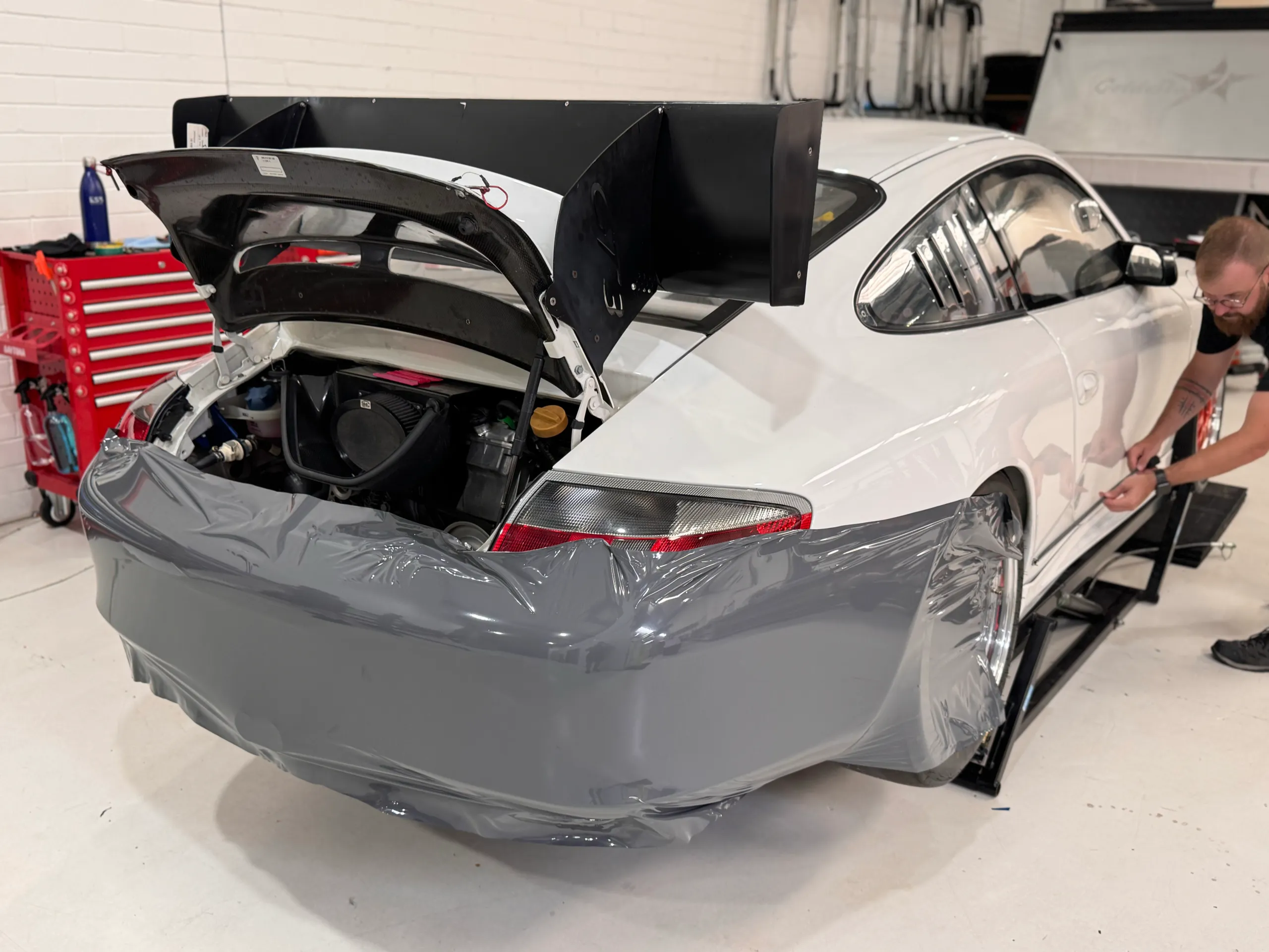 Installer fitting grey film to the Porsche rear bumper below its open engine cover and rear wing