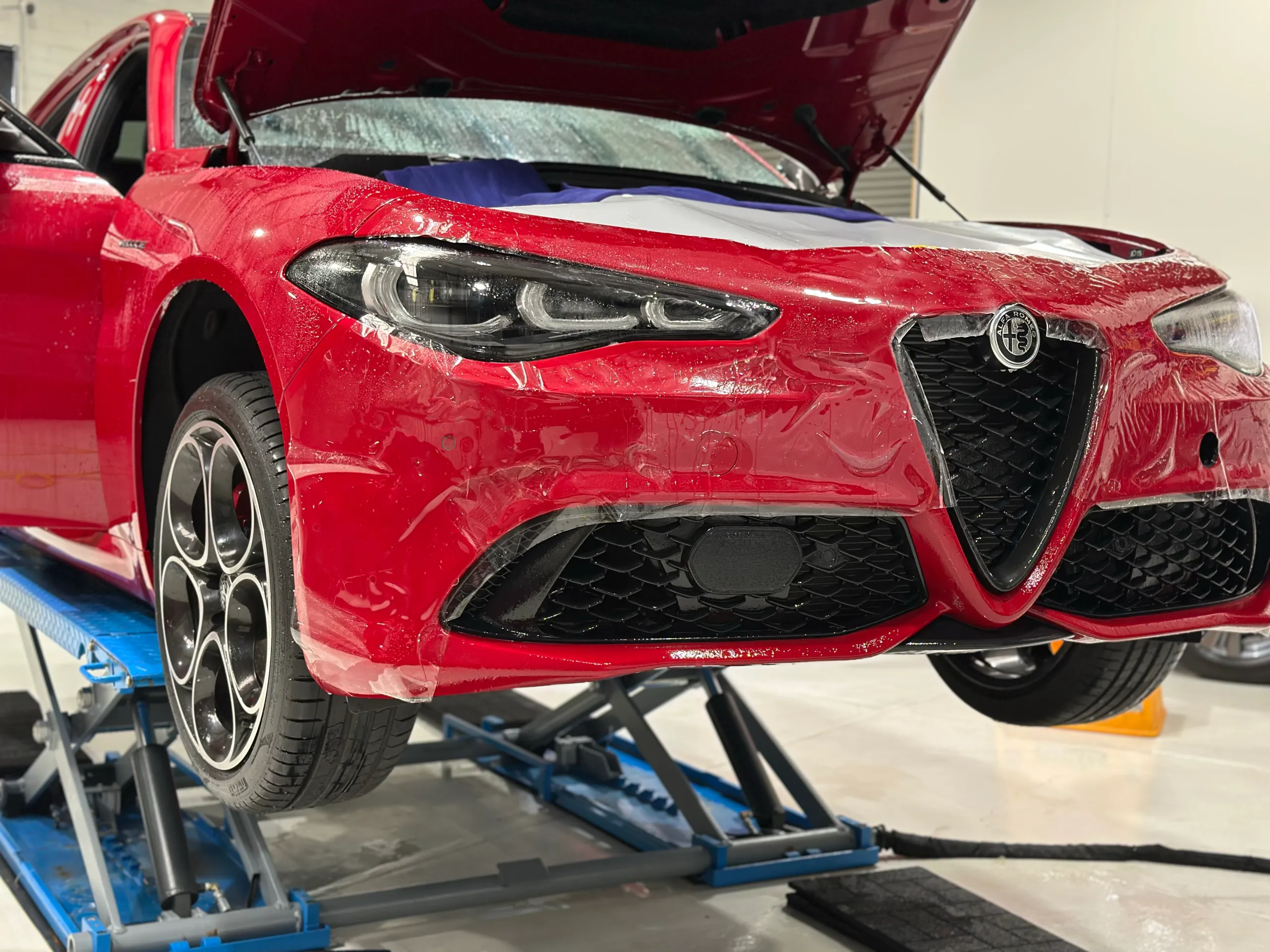 Clear film being fitted to a red Alfa Romeo Giulia front bumper on a workshop lift