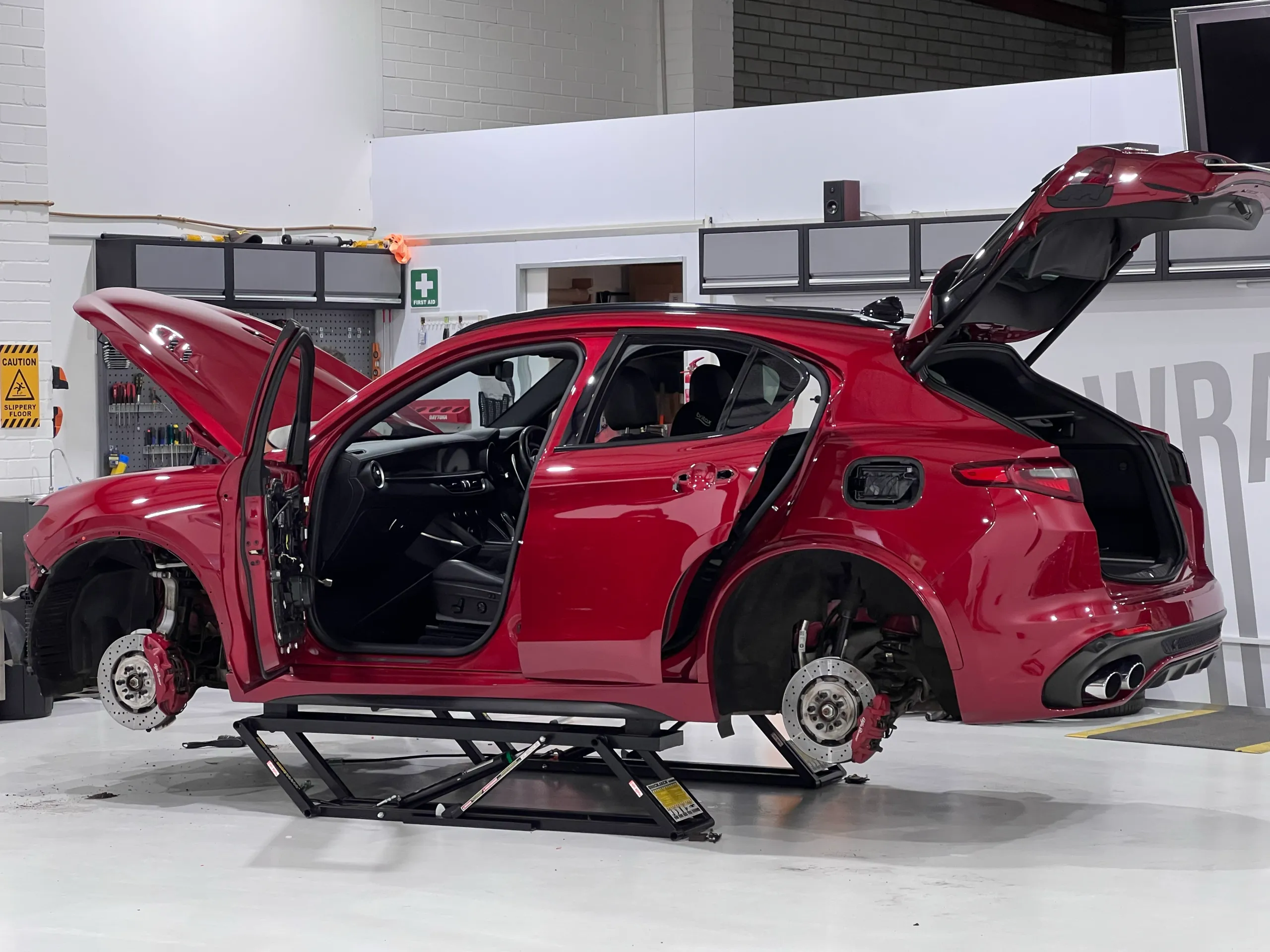 Red Alfa Romeo Stelvio on a lift with wheels and trim removed and doors open during preparation