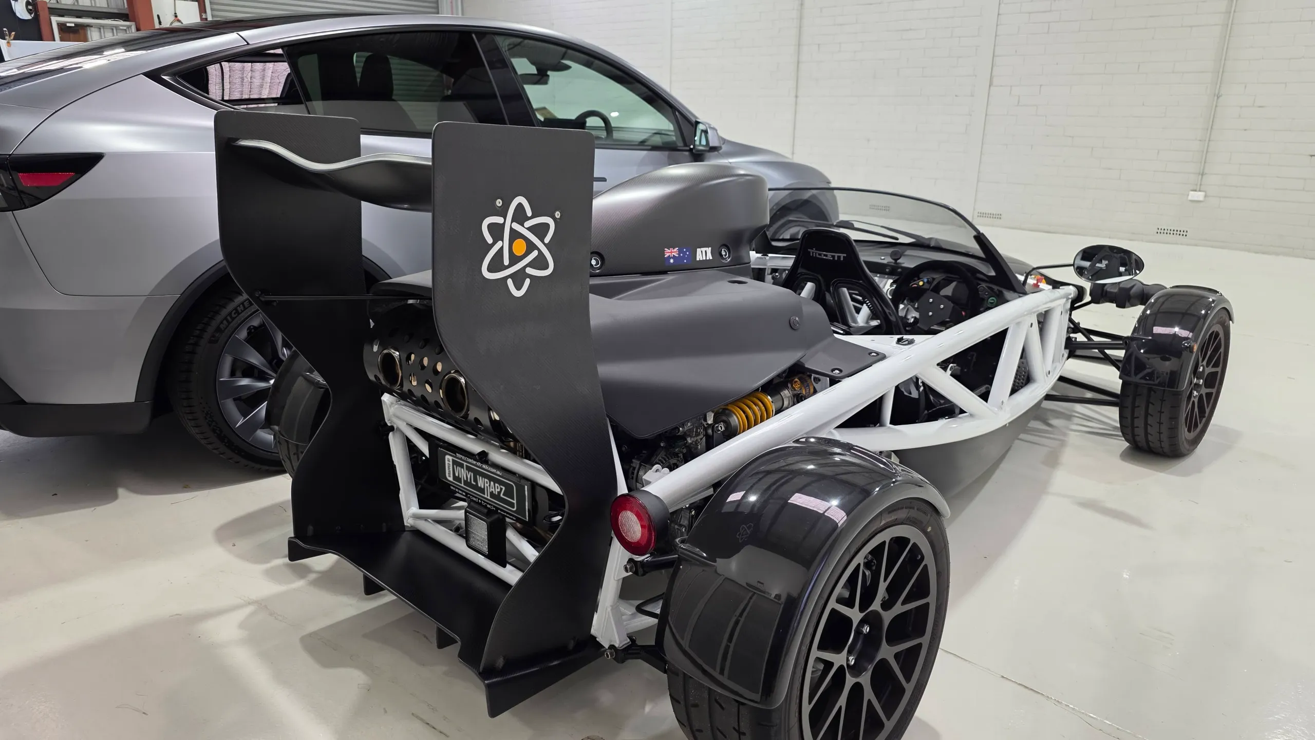 Ariel Atom rear quarter showing a tall rear wing and white tubular frame beside a Tesla