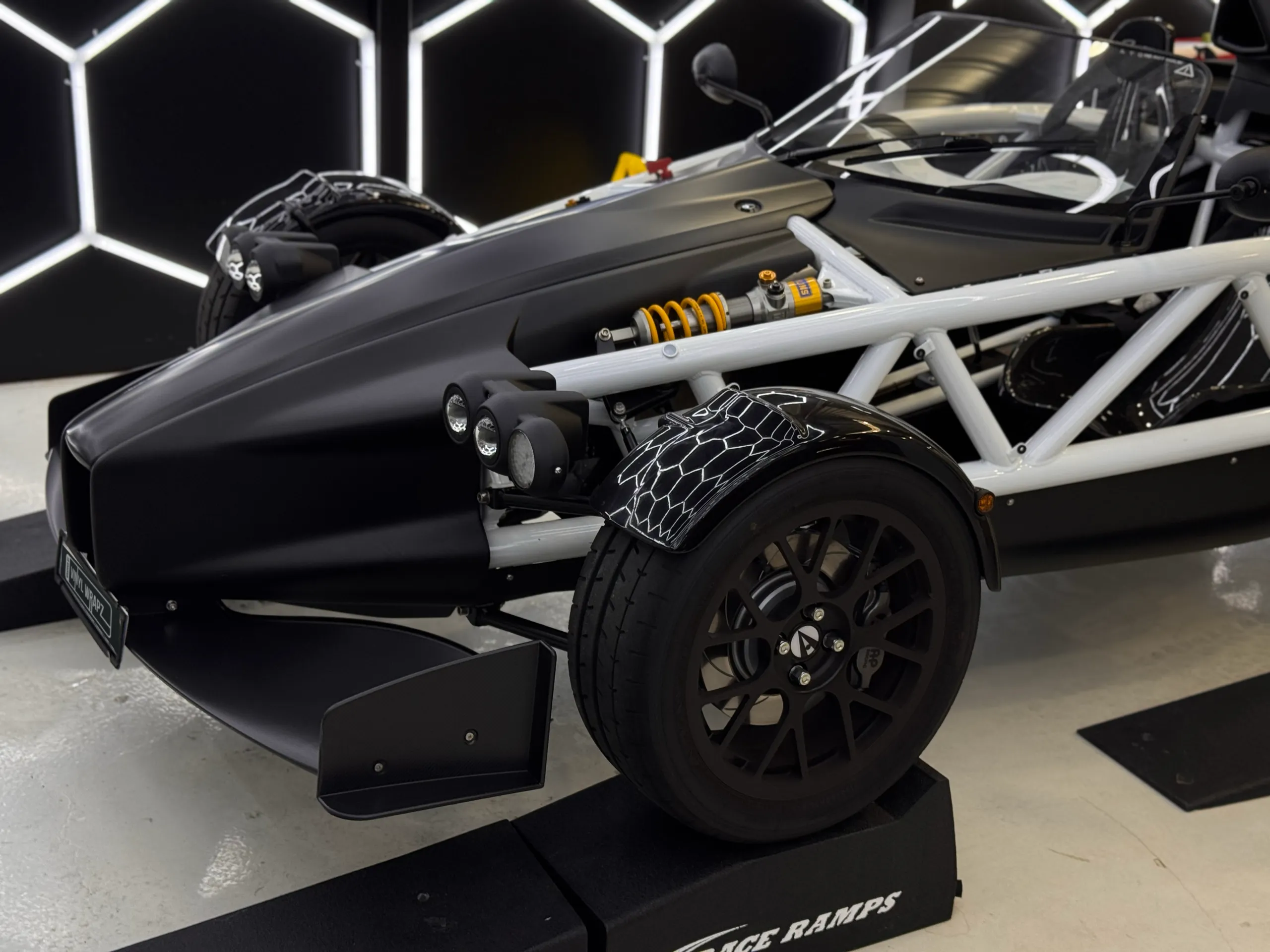 Black Ariel Atom nose and front wheel on workshop ramps beneath hexagonal lights