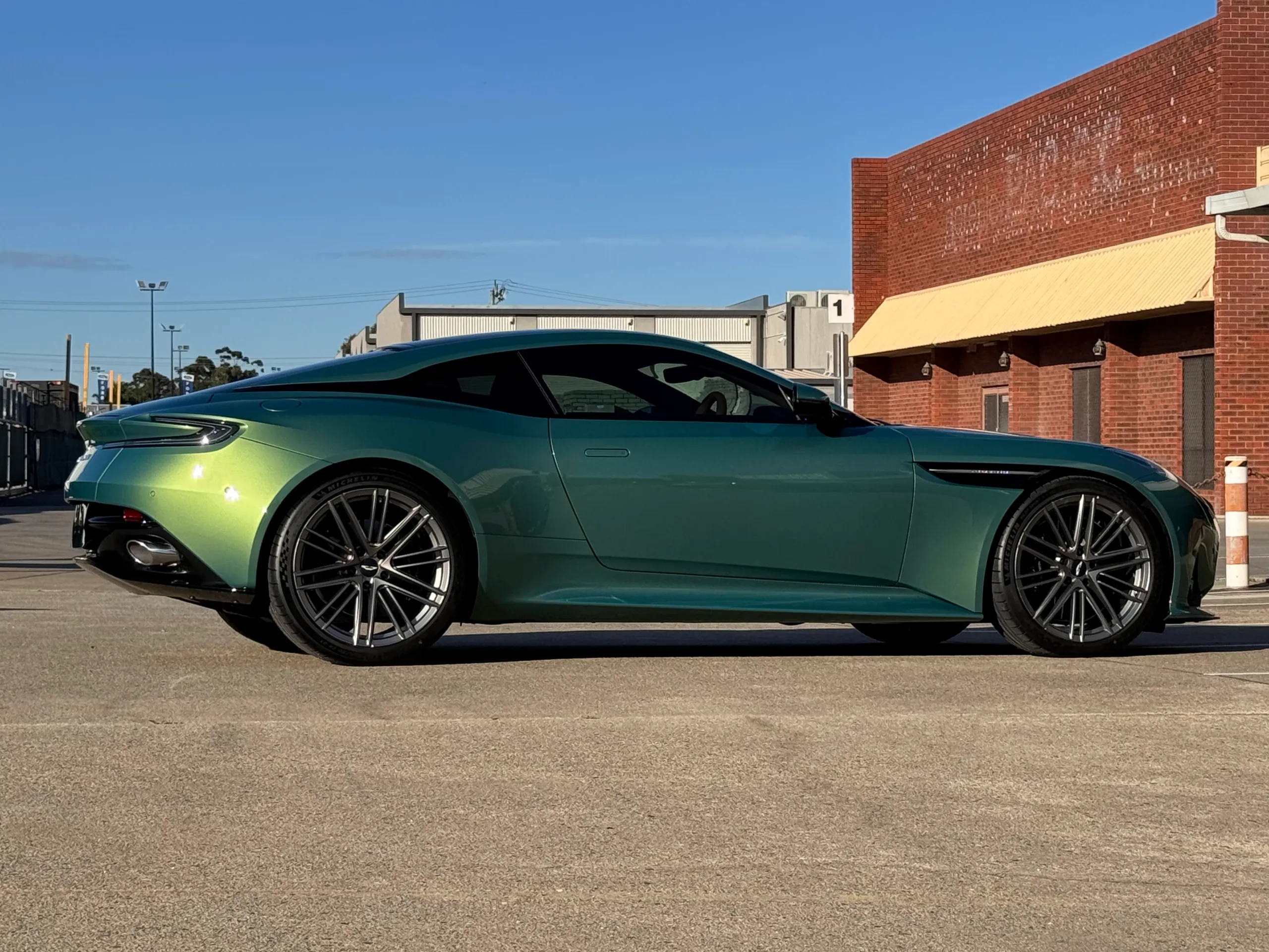 Side profile of the metallic green Aston Martin DB12 outside an industrial building