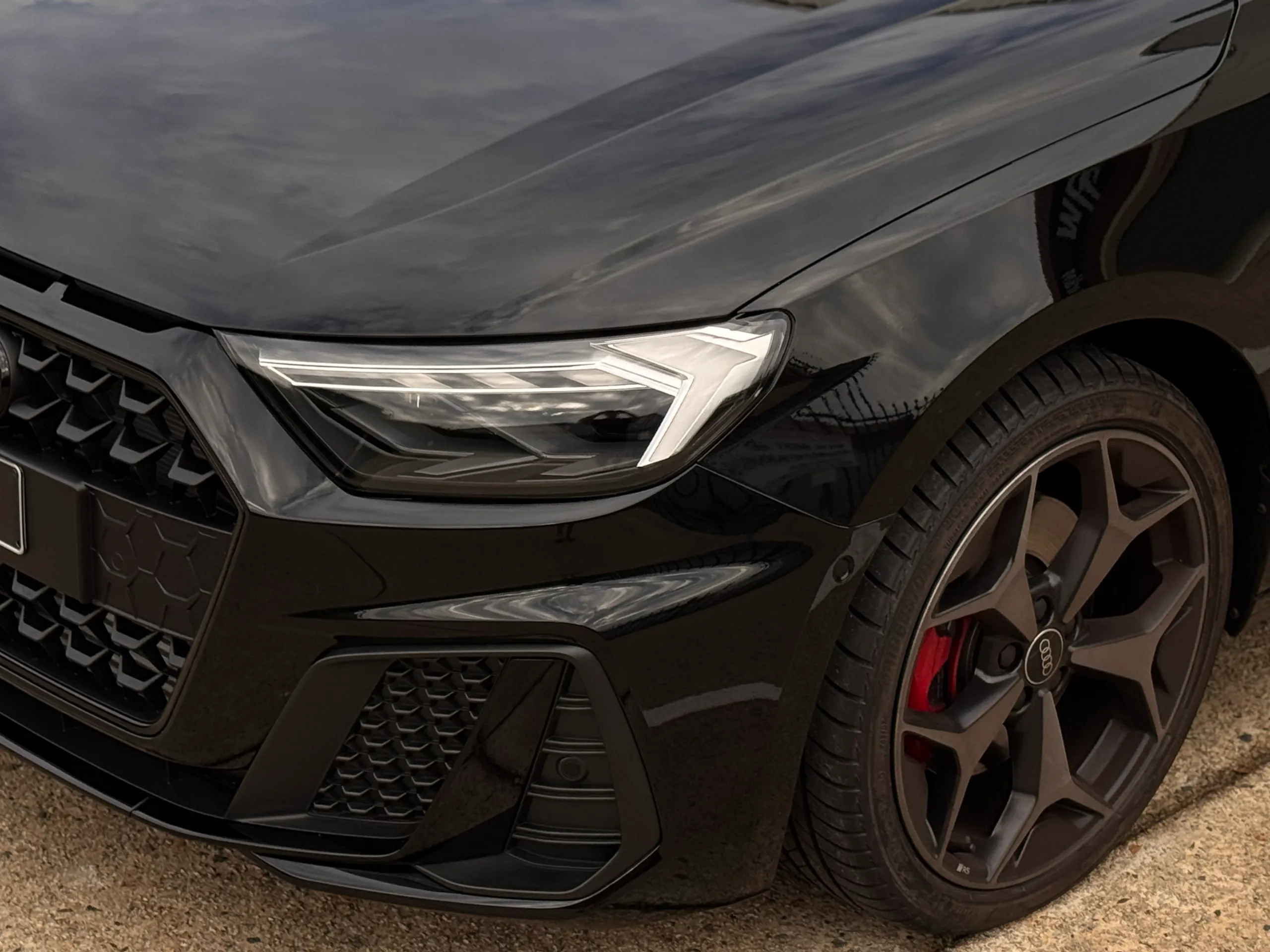 Gloss black Audi A1 headlight and front wheel outdoors, with the sky reflected in the bonnet