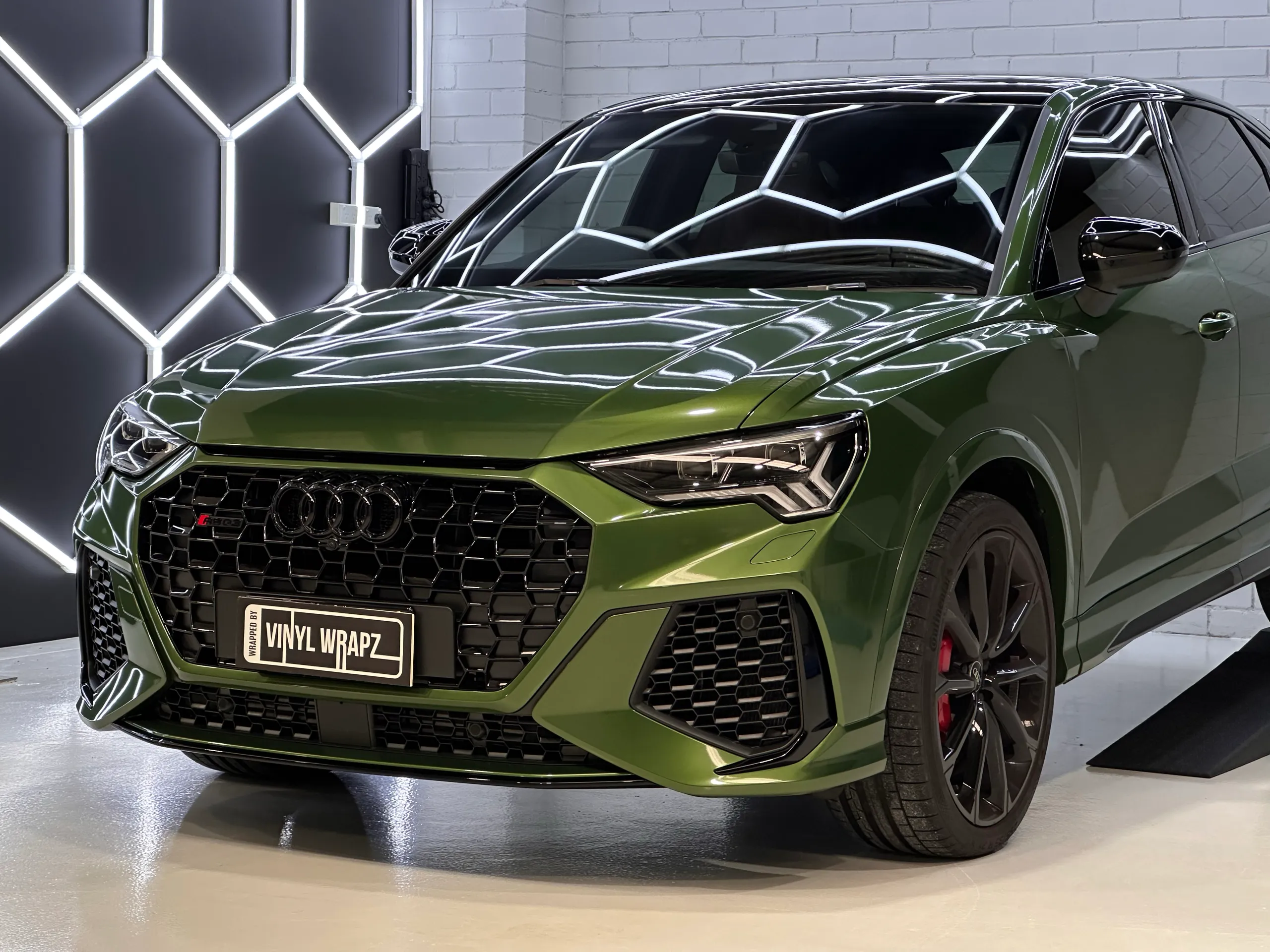 Front quarter of a metallic green Audi RS Q3 Sportback beneath hexagonal workshop lights