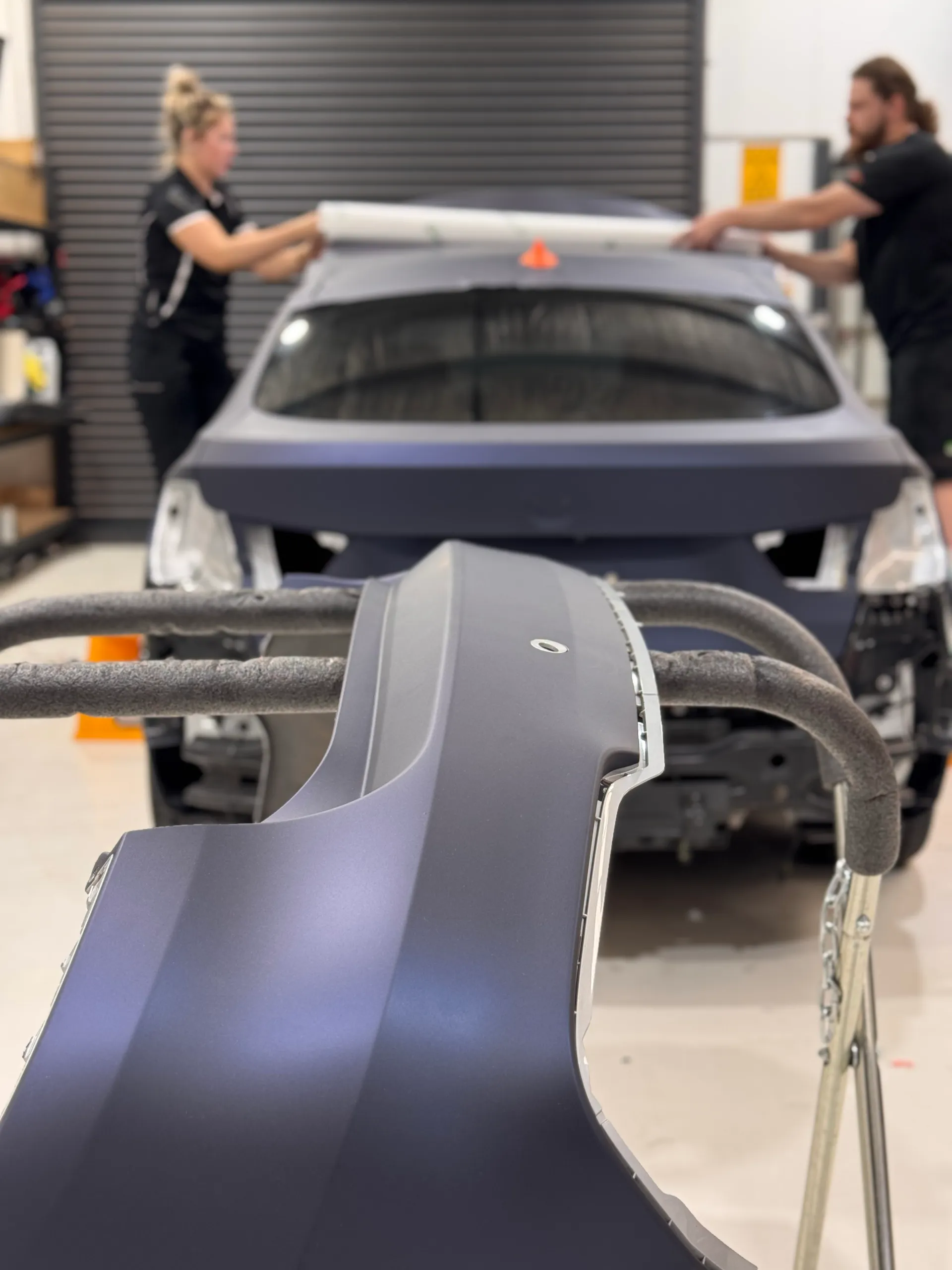 Installers fitting film to the BMW roof, with a wrapped bumper on a stand in the foreground