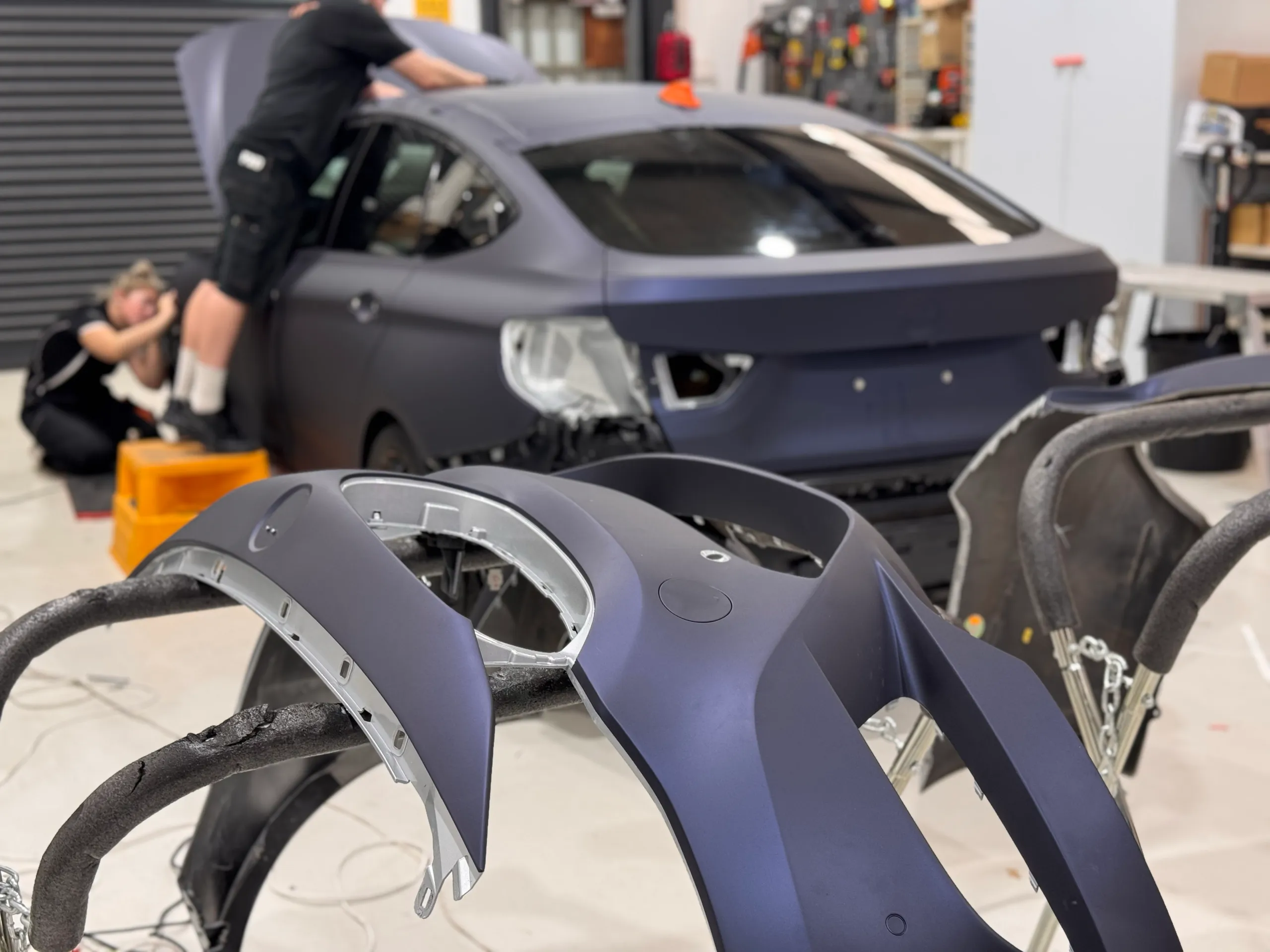 Installers wrapping the BMW Gran Turismo with removed bumper parts on stands nearby