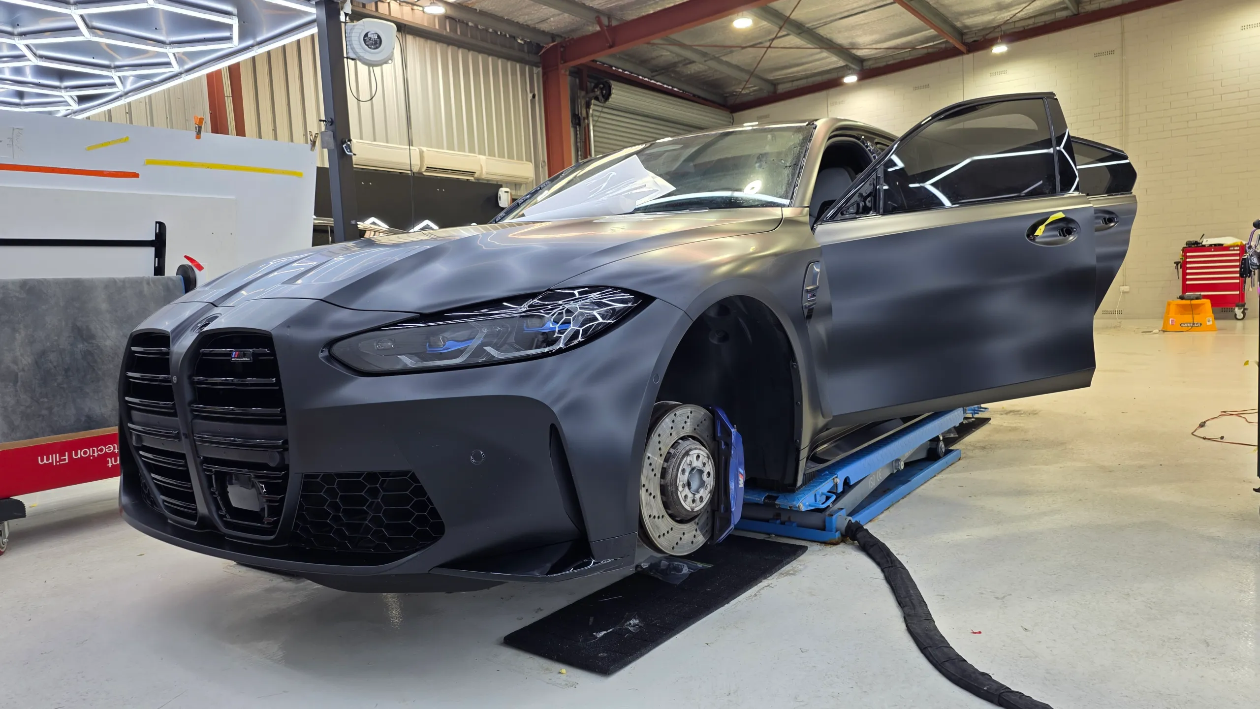 BMW M3 Competition on a workshop lift with its door open and front wheel removed during film installation
