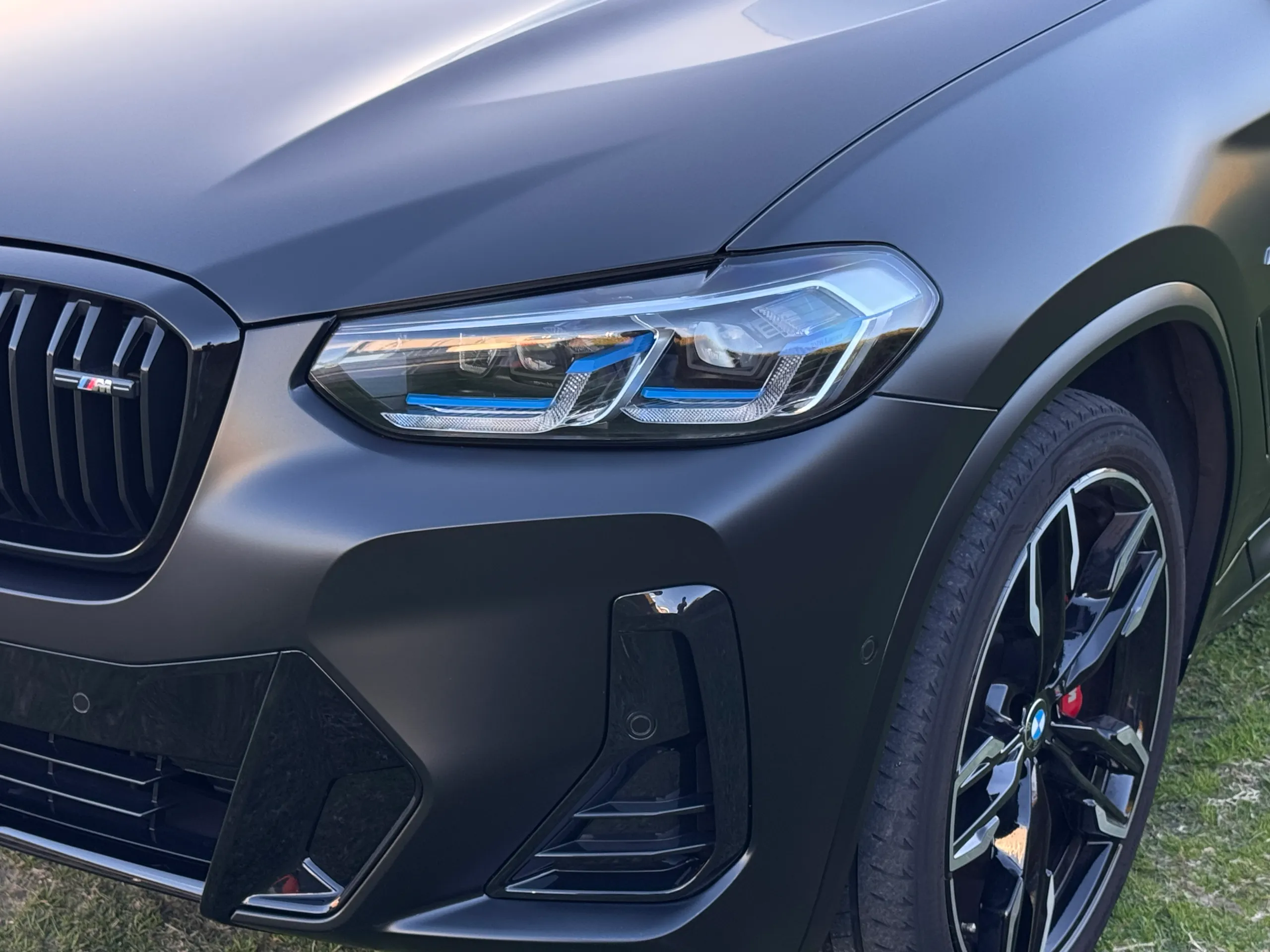 BMW X3 M40i headlight and front wheel beside matte grey bodywork and gloss black bumper trim