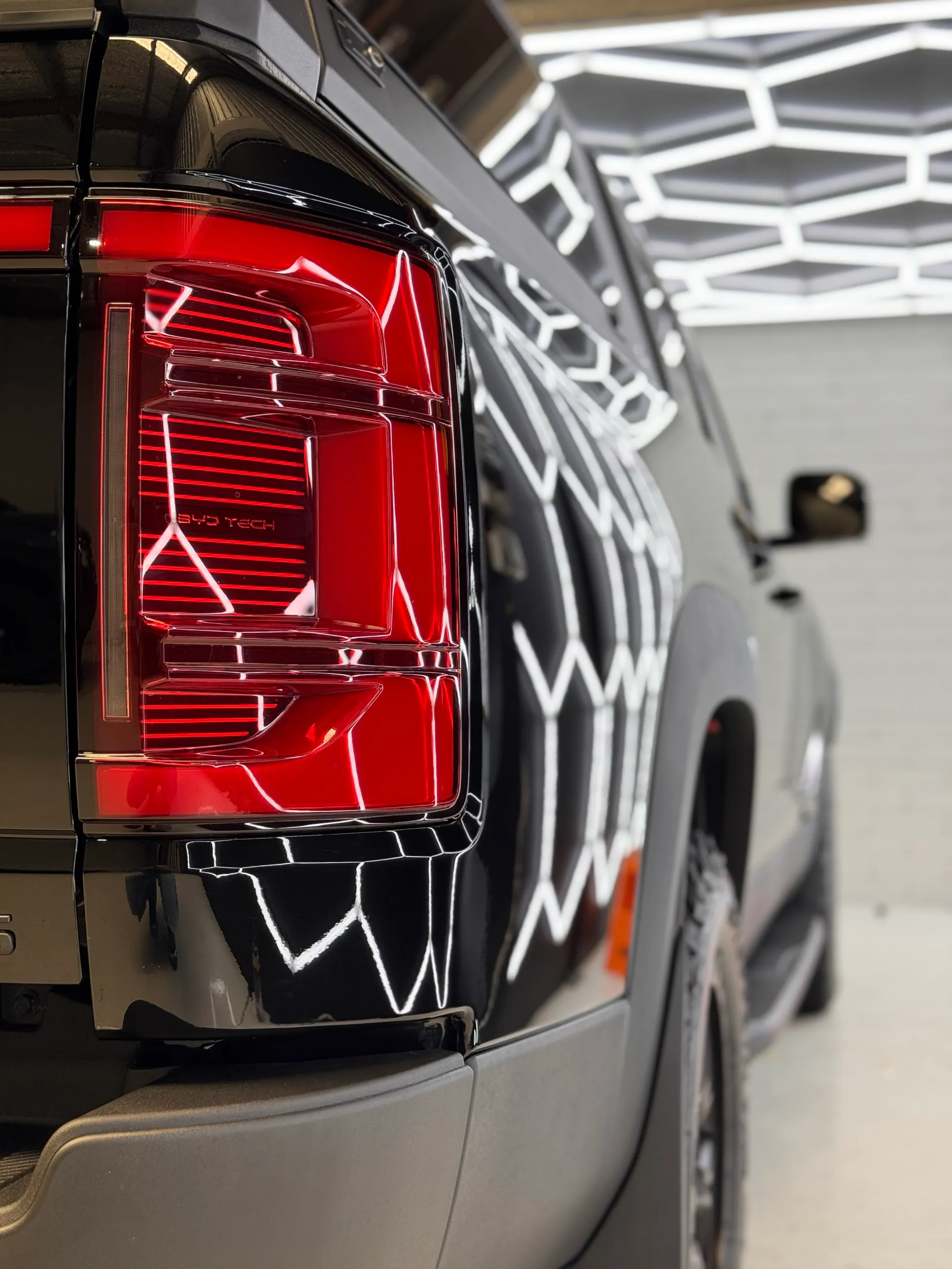 Rear light and gloss black rear quarter of the BYD Shark 6 reflecting workshop lights