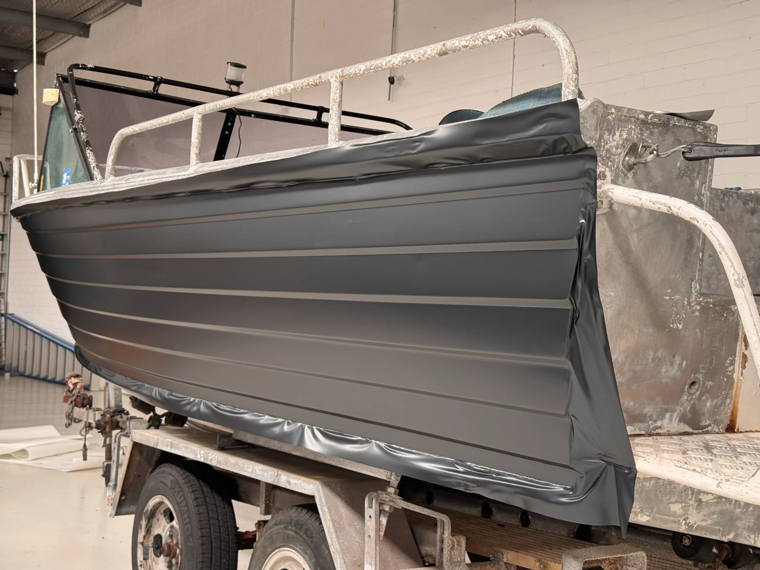Aluminium dinghy on its trailer with grey film fitted along one side and excess film around the edges
