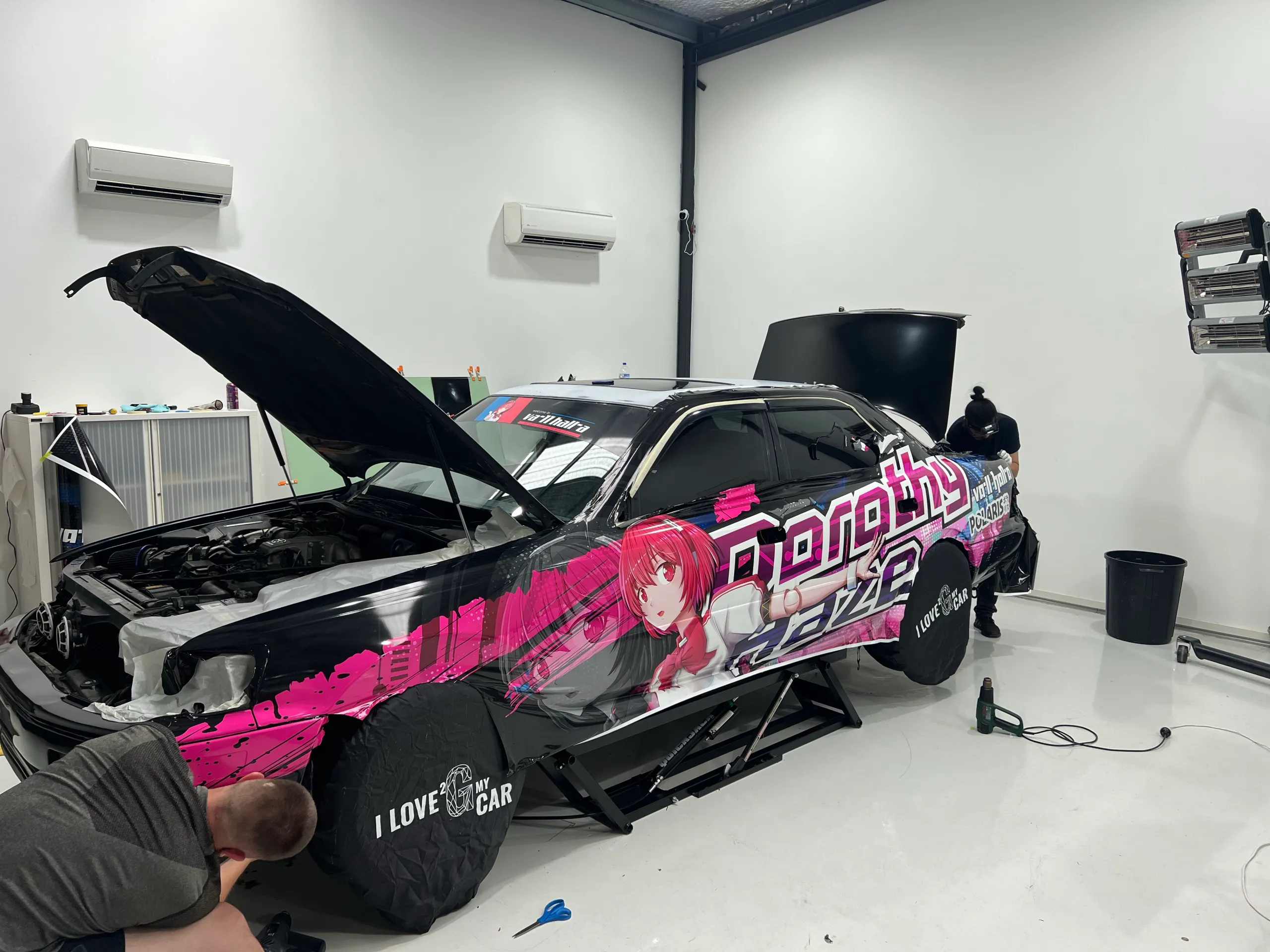 Installers kneeling beside the Toyota while fitting anime graphics