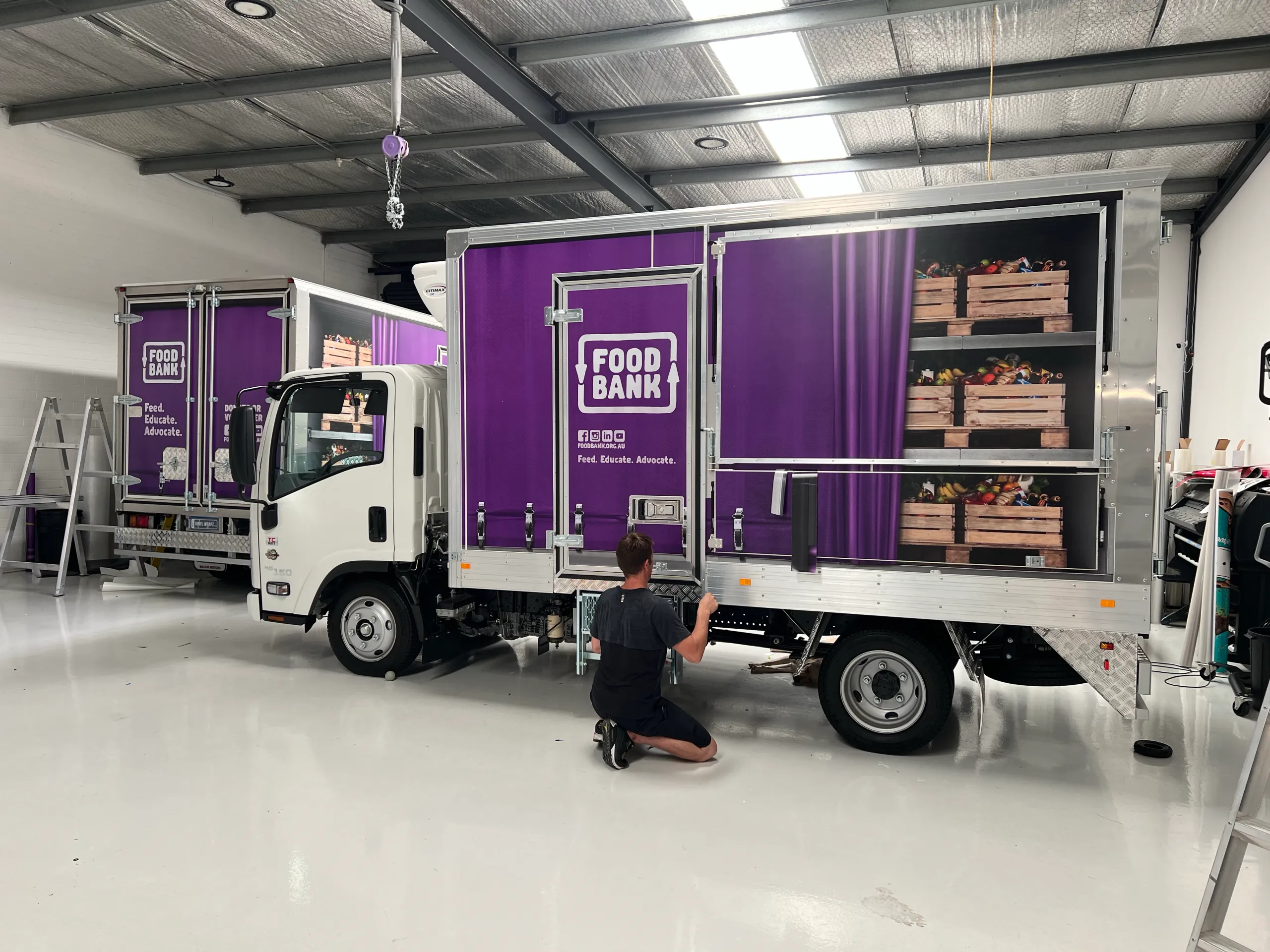 Back-facing installer working on a Foodbank truck with purple graphics and printed produce shelves