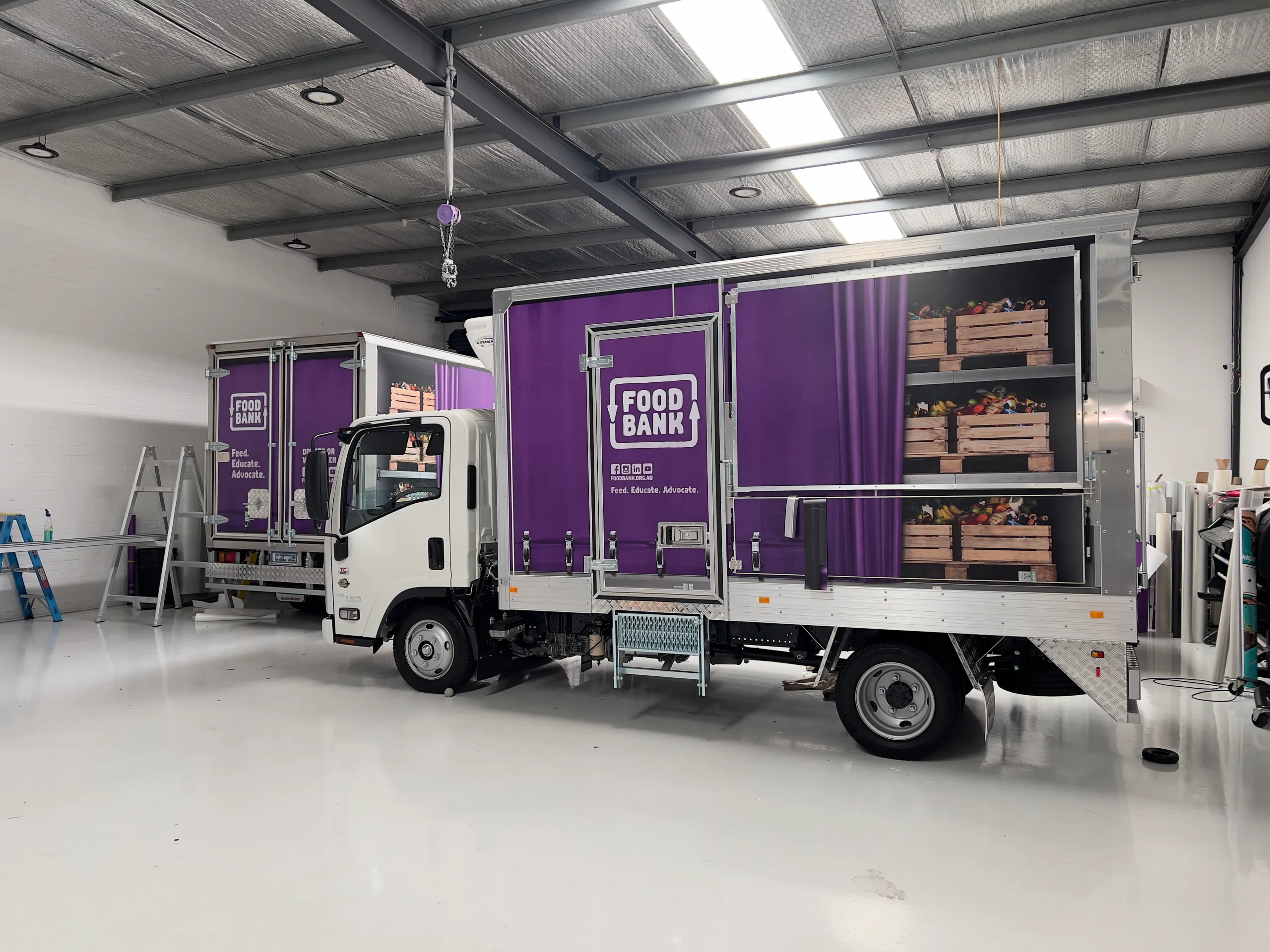 Two Foodbank trucks inside the workshop, showing purple branding and printed produce shelves