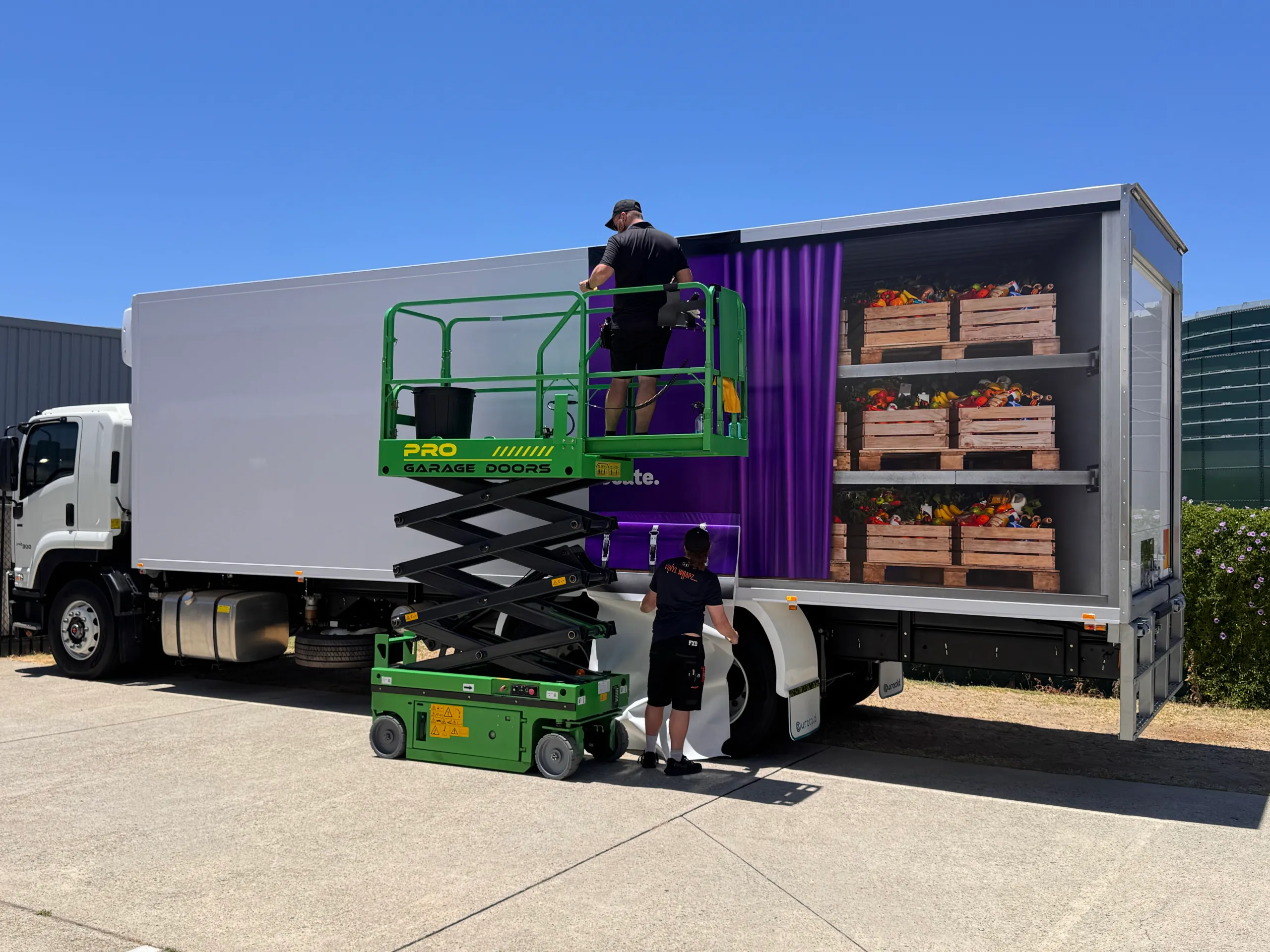 Installers fitting purple Foodbank graphics to a refrigerated truck using a scissor lift