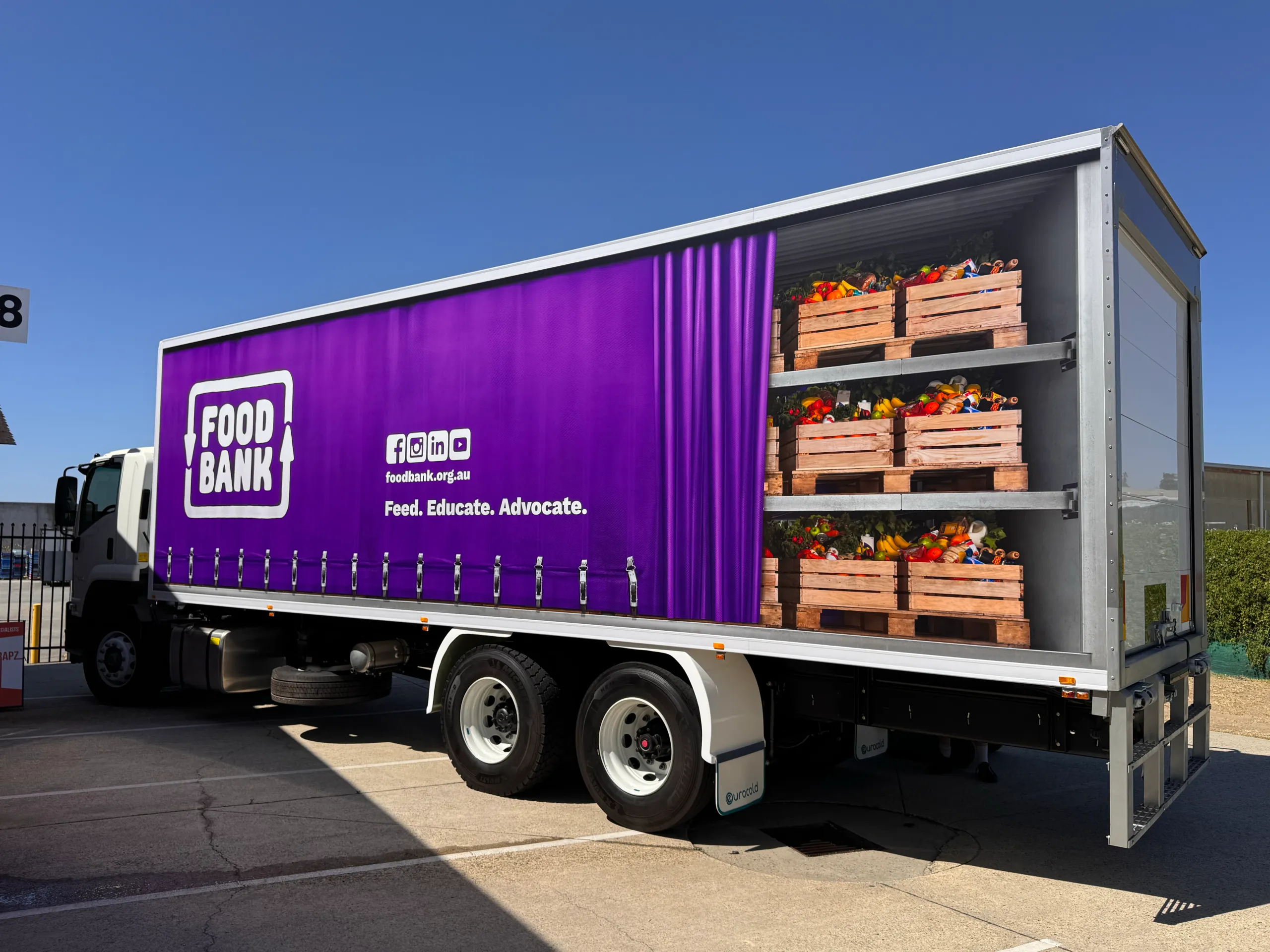 Foodbank truck with purple curtain and produce-crate graphics across its side