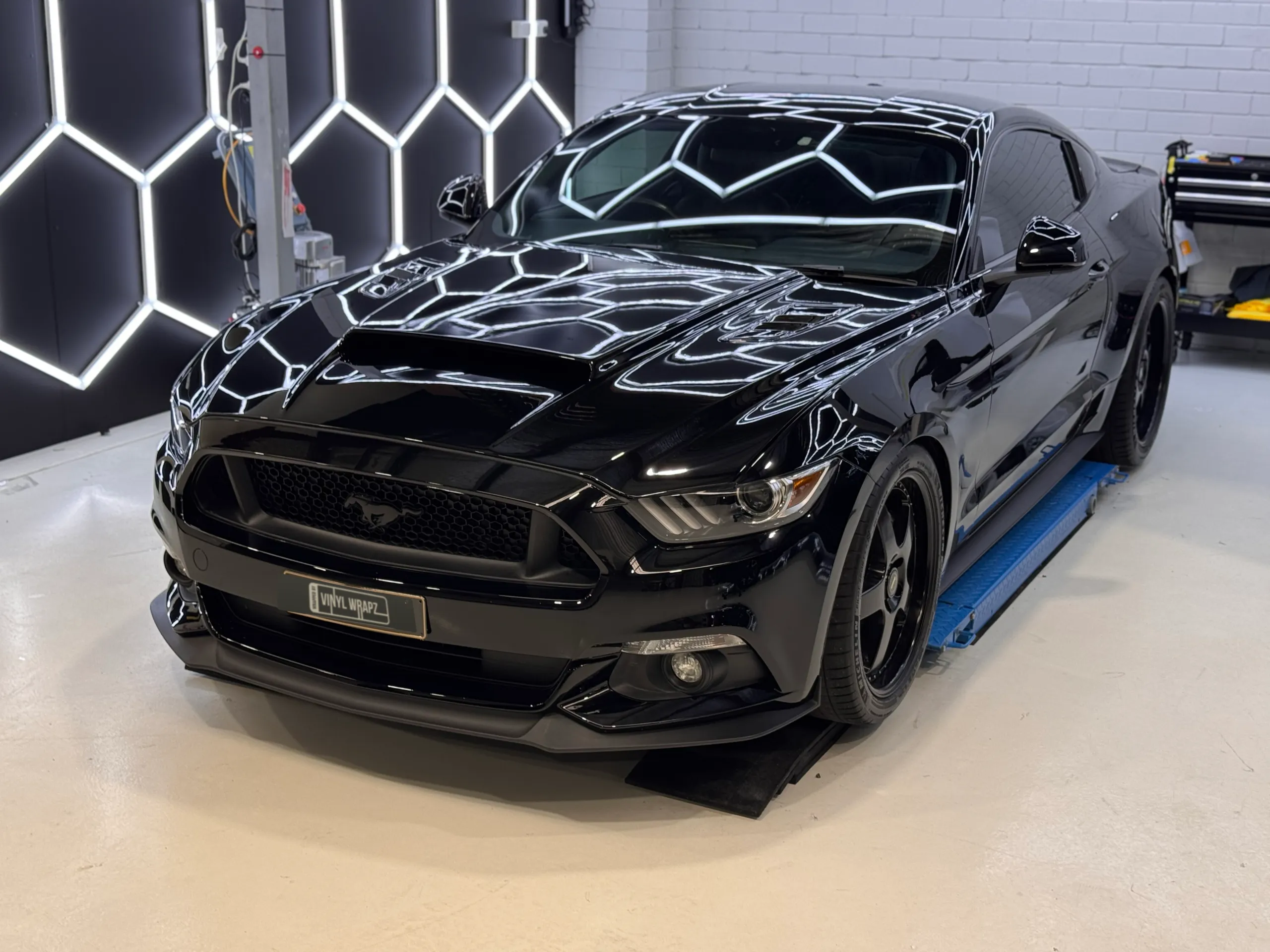 Gloss black Ford Mustang on a workshop lift beneath hexagonal lights