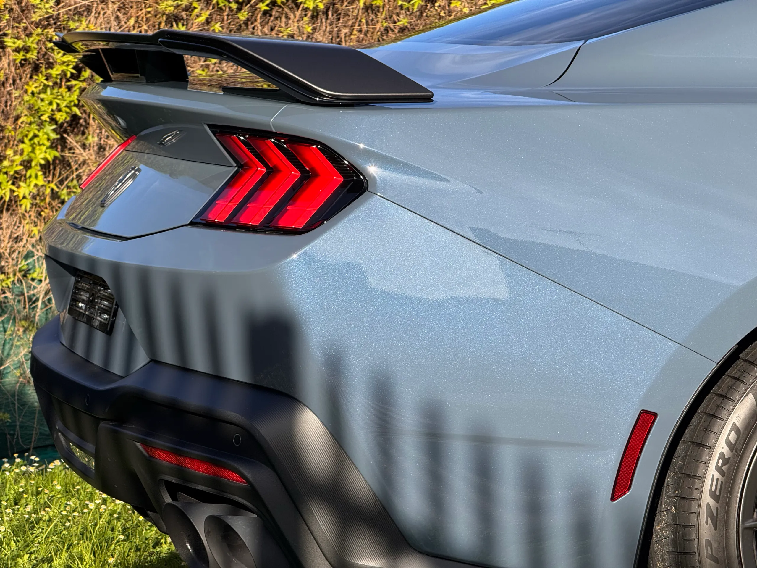 Blue-grey Ford Mustang Dark Horse rear quarter in sunlight with fence shadows across the bumper