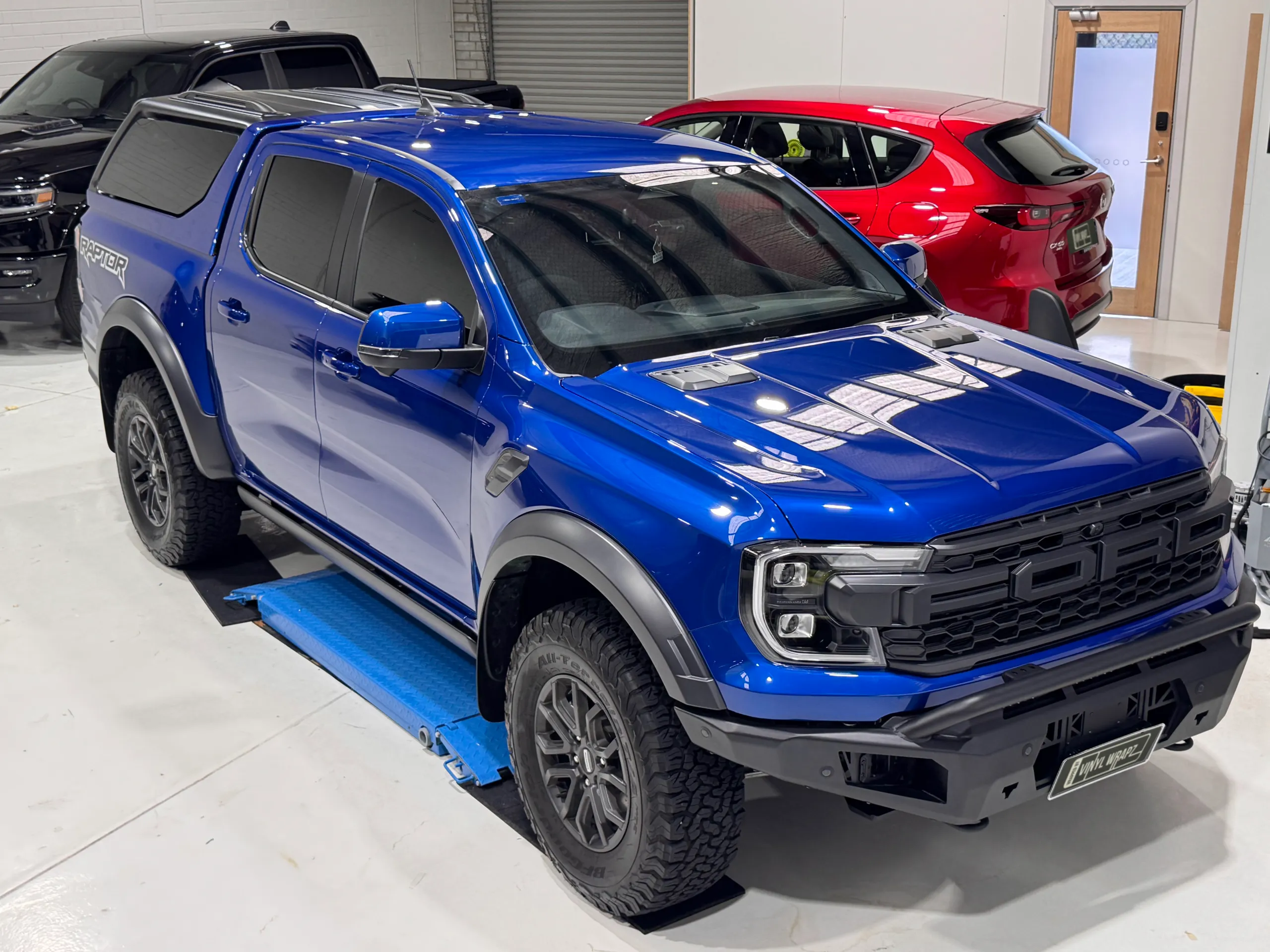Elevated front-quarter view of a gloss blue Ford Ranger Raptor with a canopy and black front bar