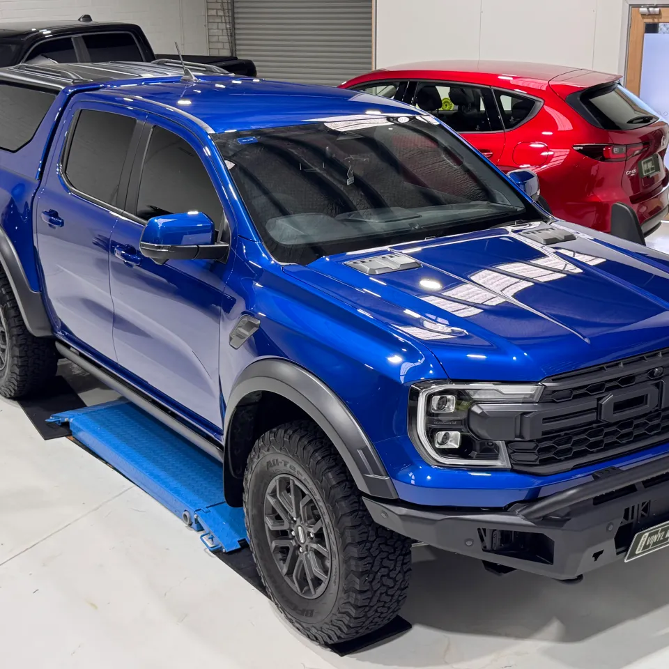 Elevated front-quarter view of a gloss blue Ford Ranger Raptor with a canopy and black front bar