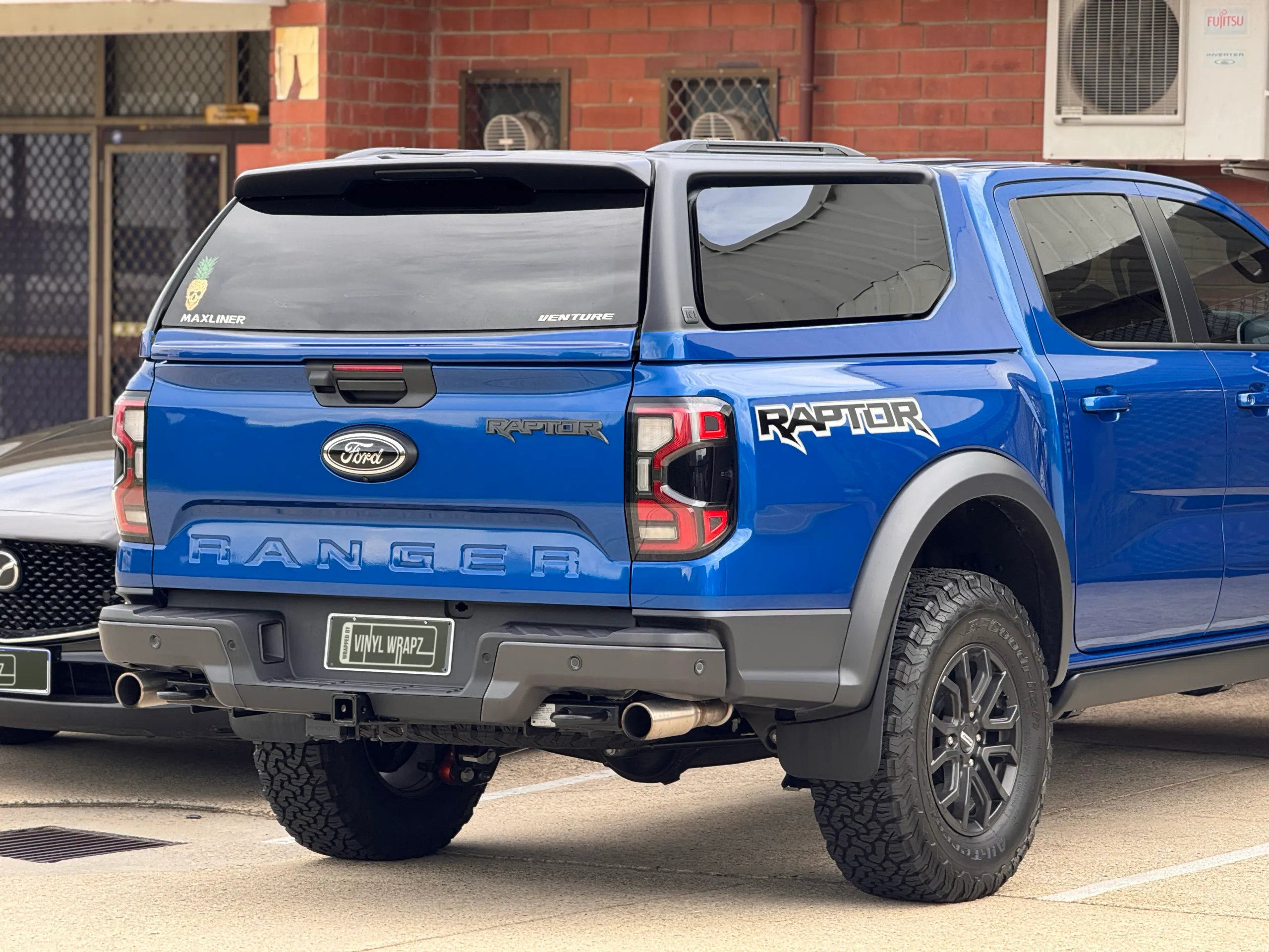 Rear-quarter view of the gloss blue Ford Ranger Raptor outdoors with another car behind it