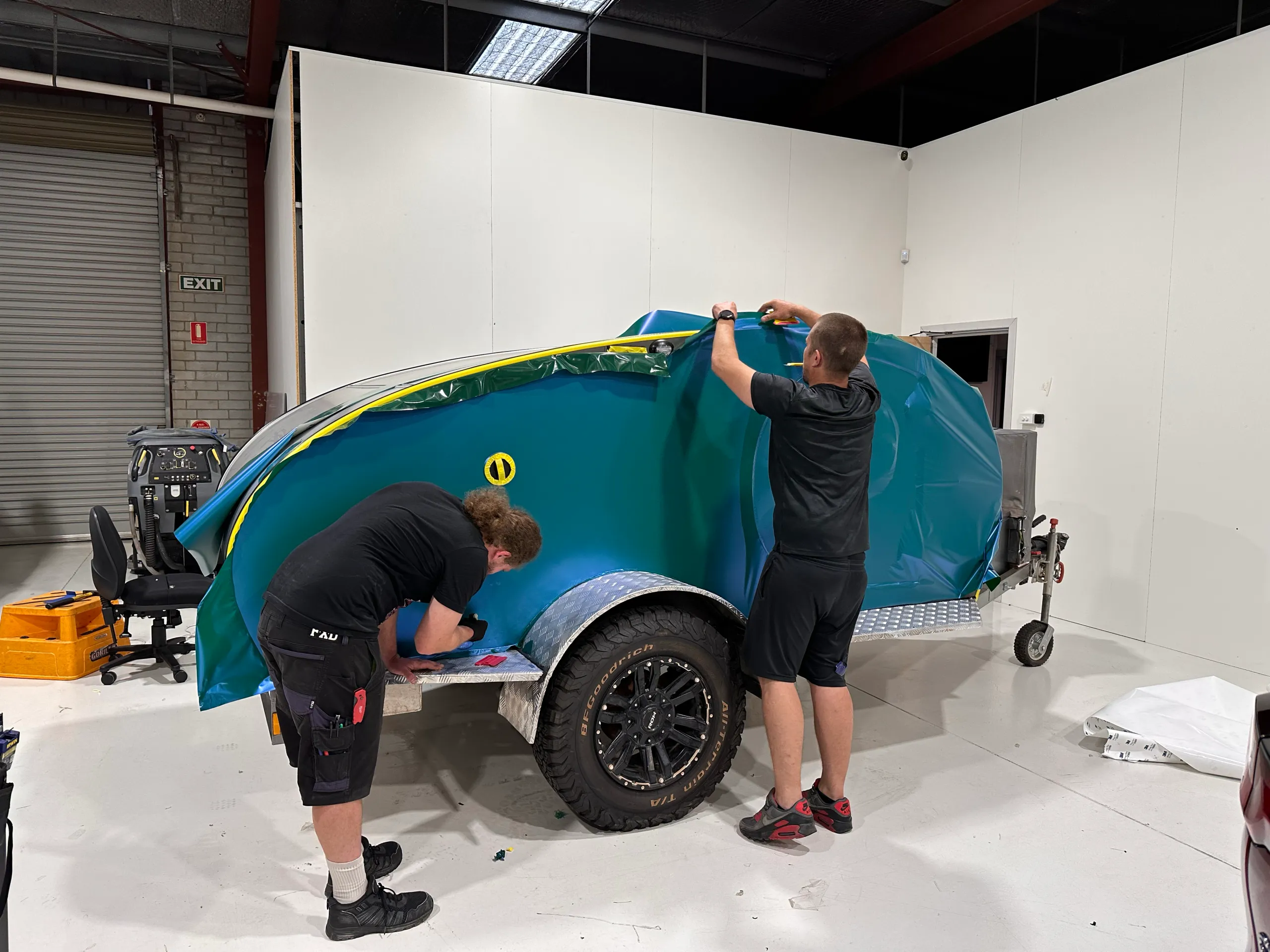 Installers fitting green and blue colour-shift film to a camping trailer