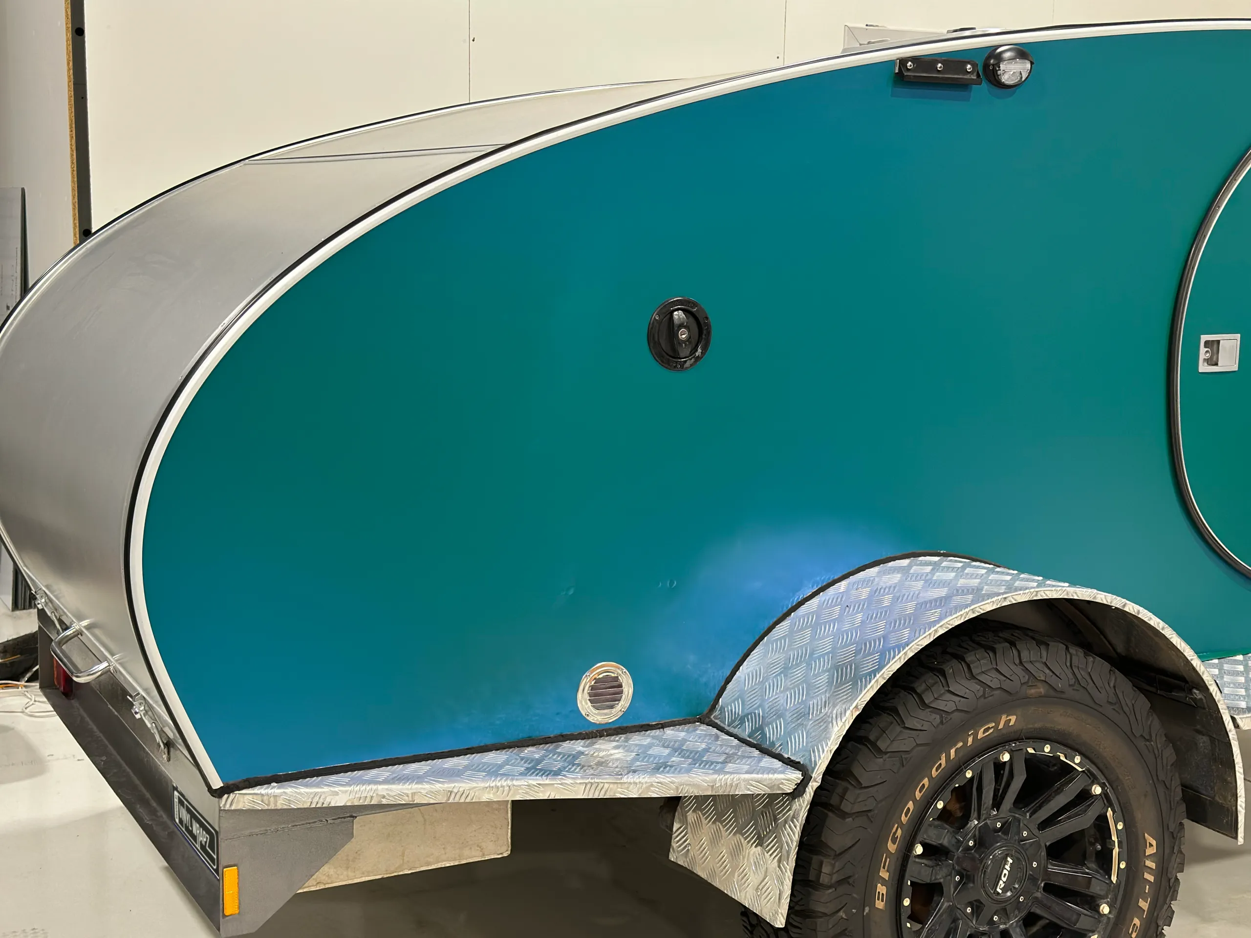 Iridescent green camping trailer side above a black wheel and checkerplate trim