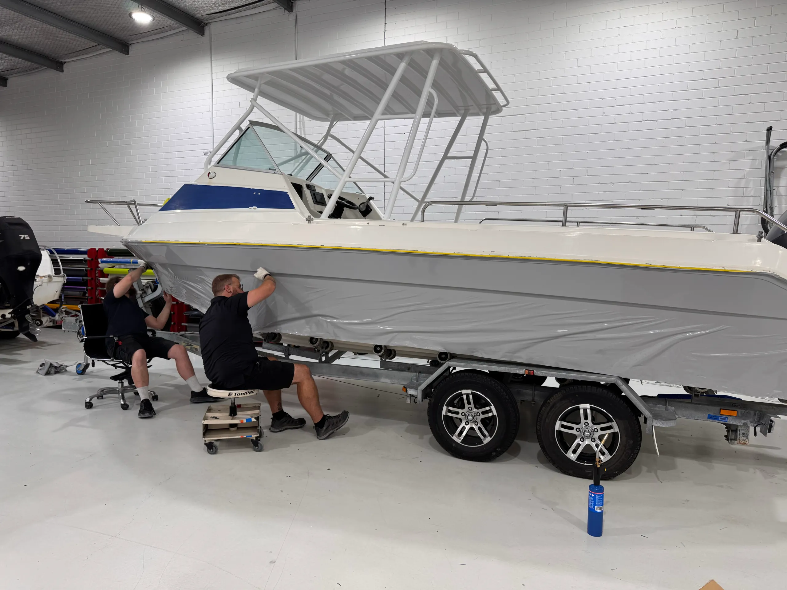 Installers fitting grey film along a white boat hull on its trailer in the workshop