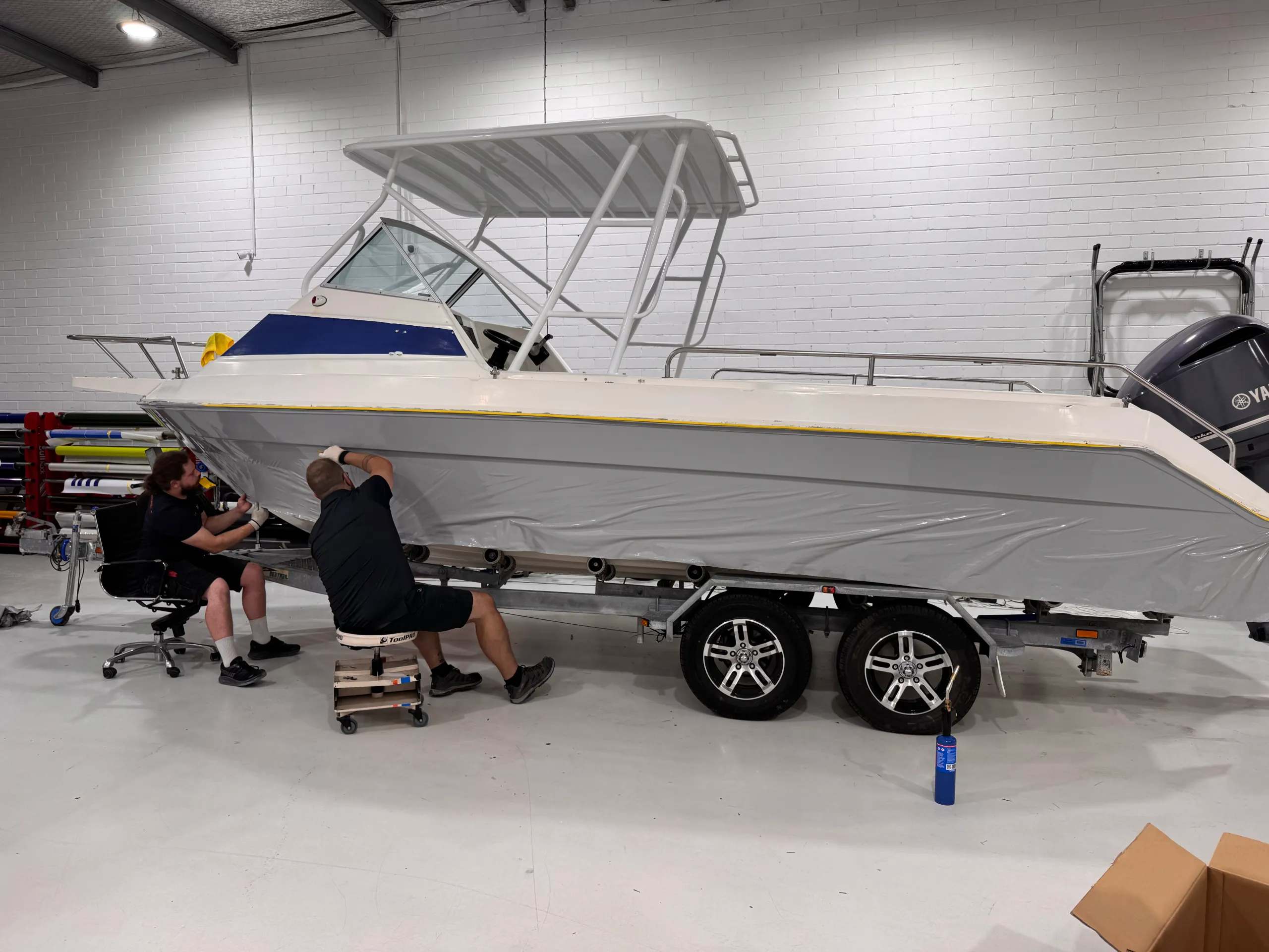 Grey film being fitted along the boat hull beneath its white cabin and canopy