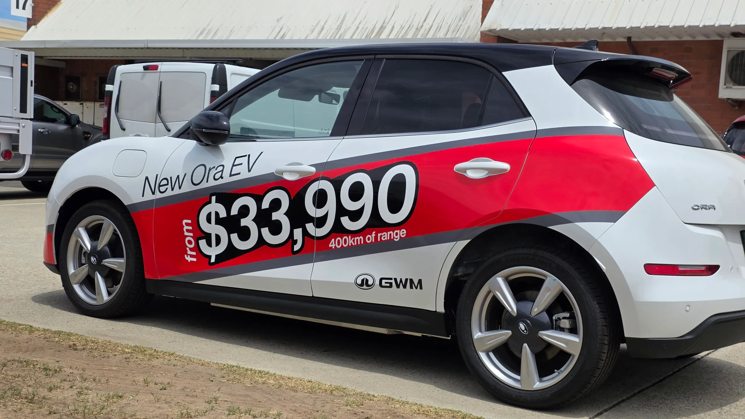 Rear-quarter view of a white GWM Ora with red and grey promotional graphics across its doors
