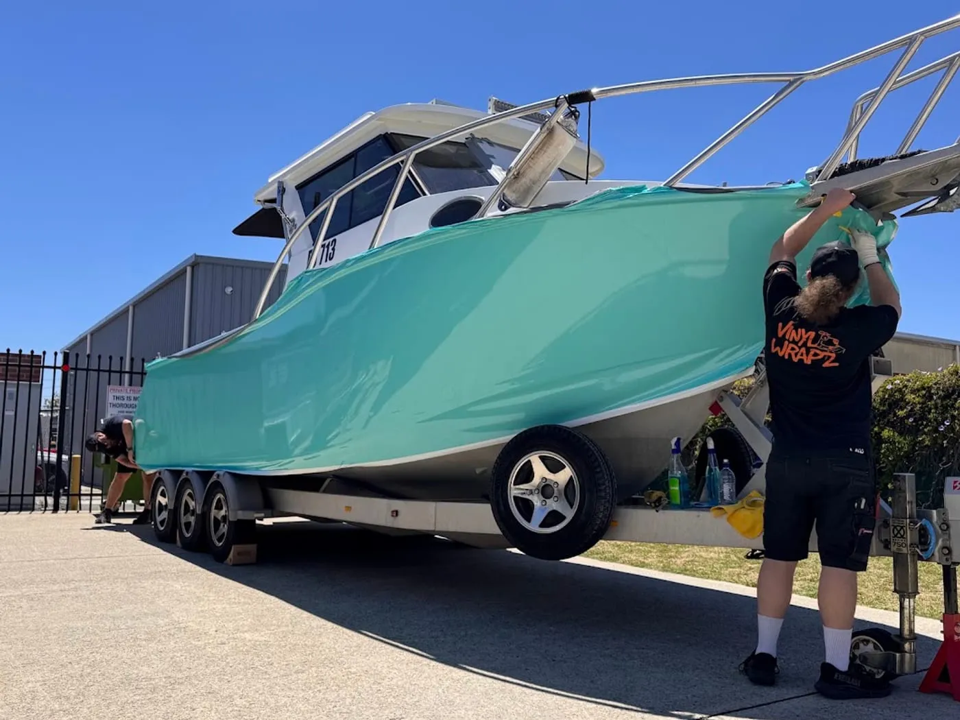 Installer fitting gloss teal wrap to a cabin cruiser hull outside the workshop