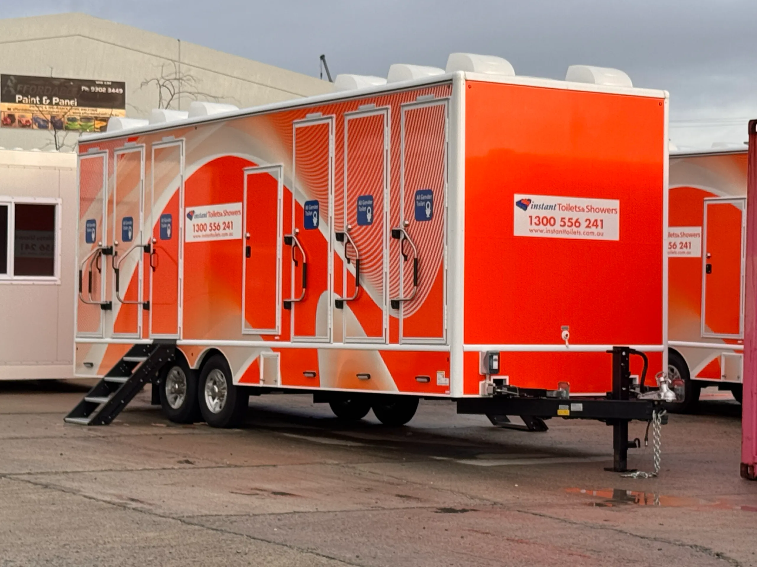 Orange and white Instant Toilets & Showers trailer parked outdoors with multiple doors and access steps