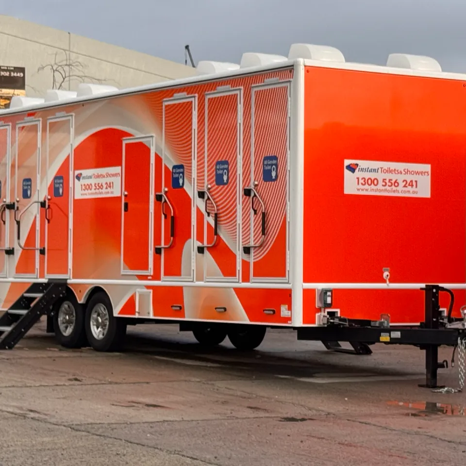 Orange and white Instant Toilets & Showers trailer parked outdoors with multiple doors and access steps