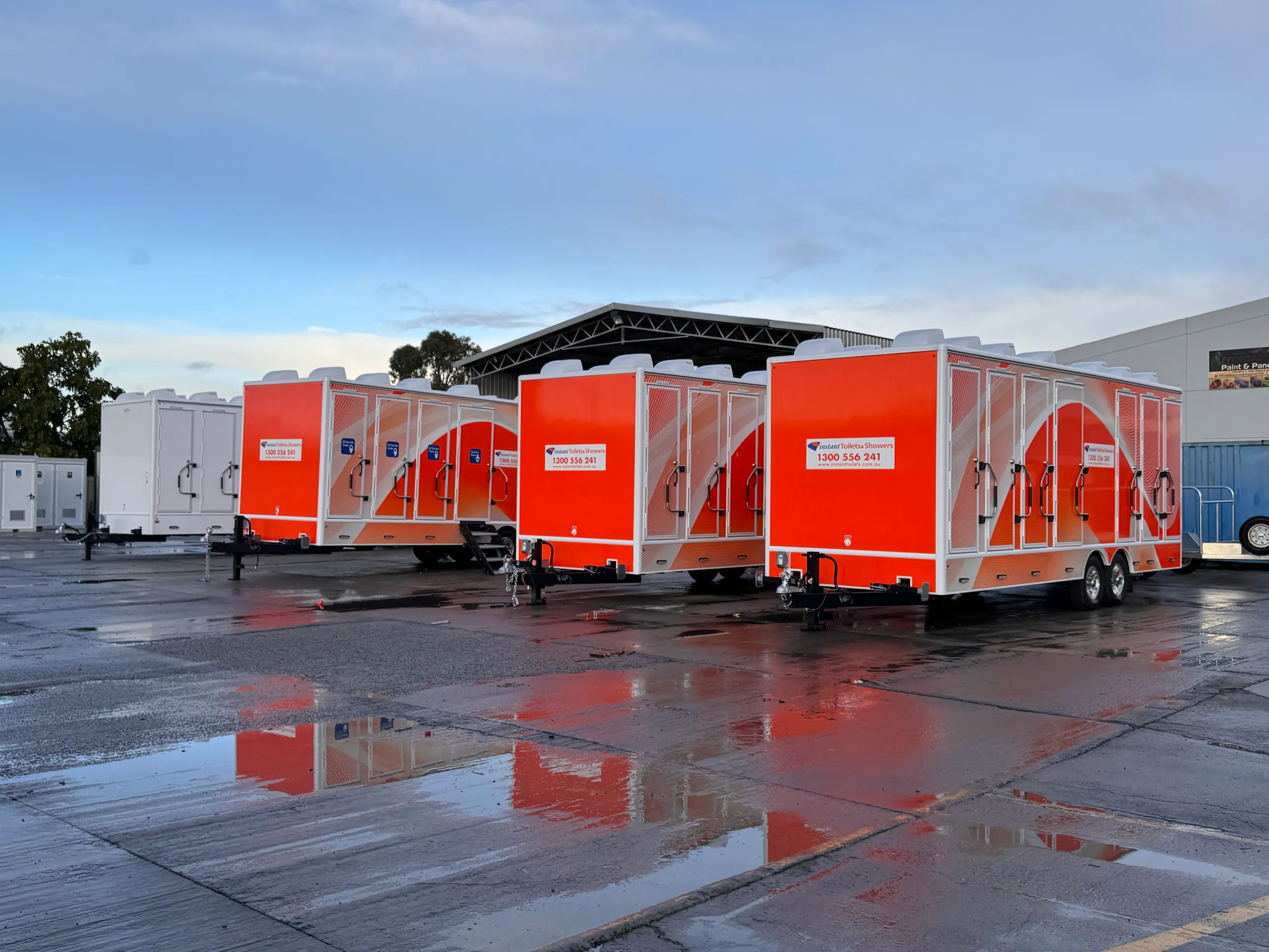 Fleet of orange and white Instant Toilets & Showers trailers reflected in a wet yard