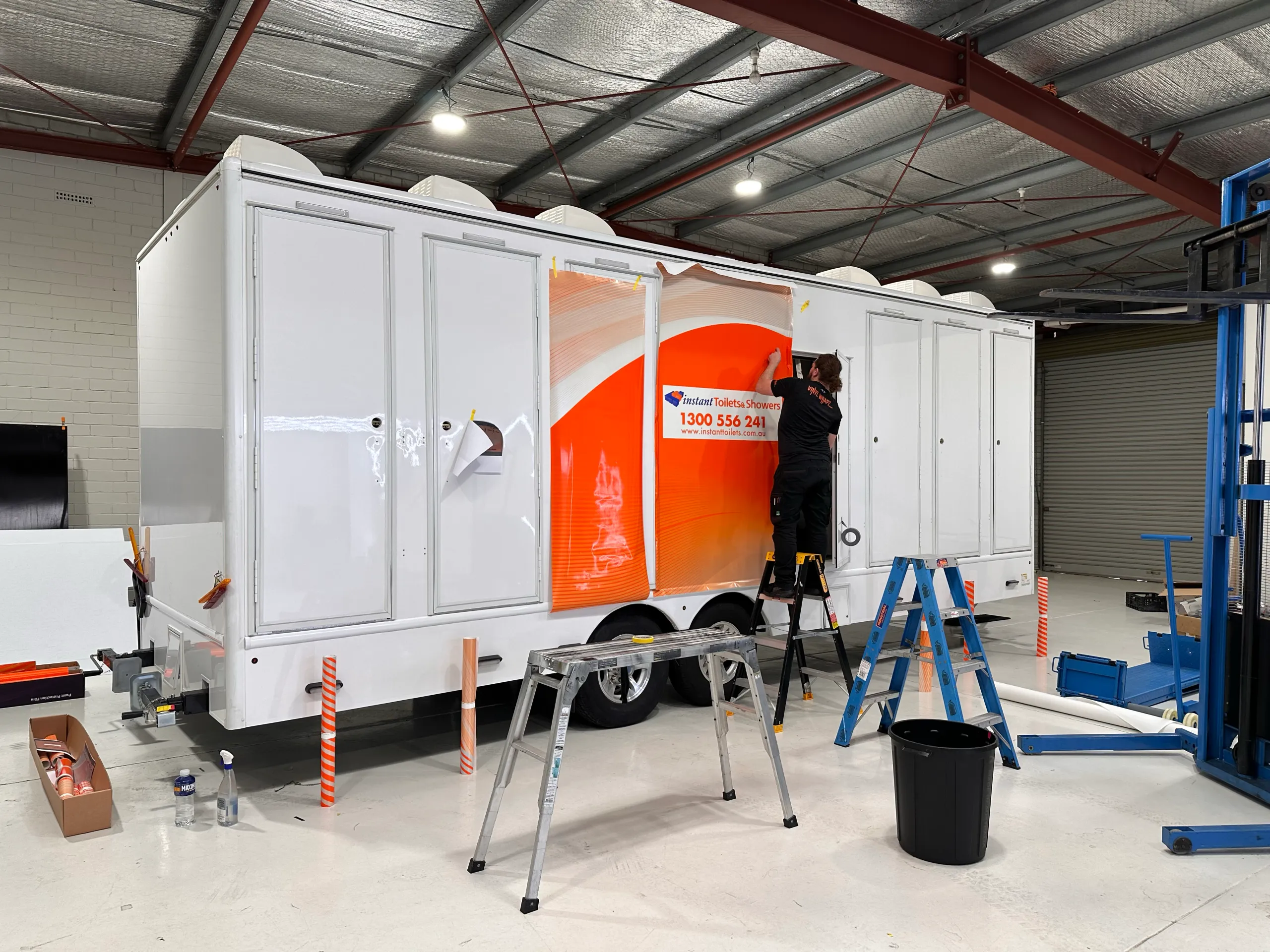 Back-facing installer fitting orange graphics to a white amenities trailer inside the workshop