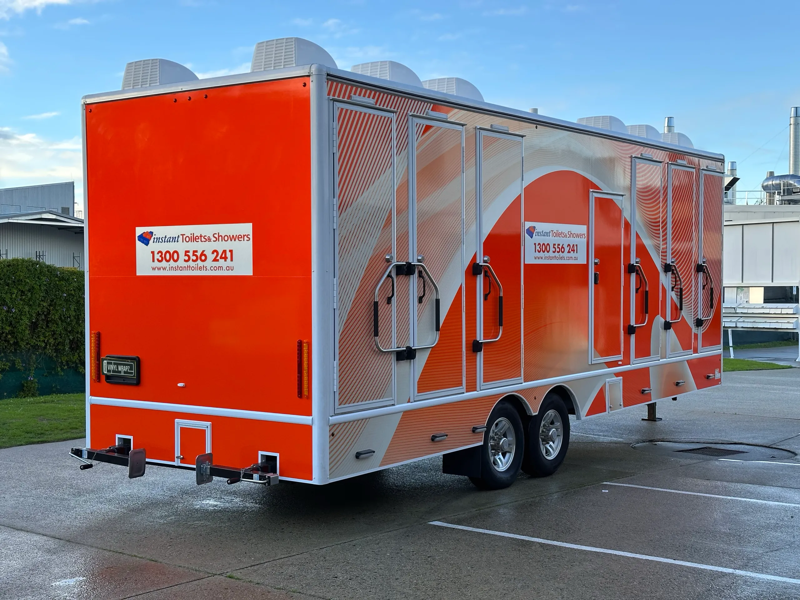 Orange and white Instant Toilets & Showers trailer viewed from the rear quarter outside the workshop