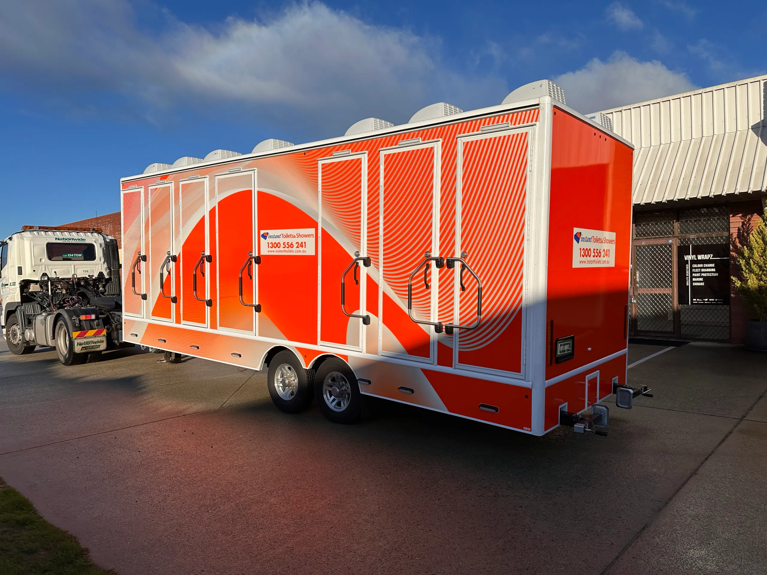 Wrapped Instant Toilets & Showers trailer outside the workshop, with orange graphics across its doors