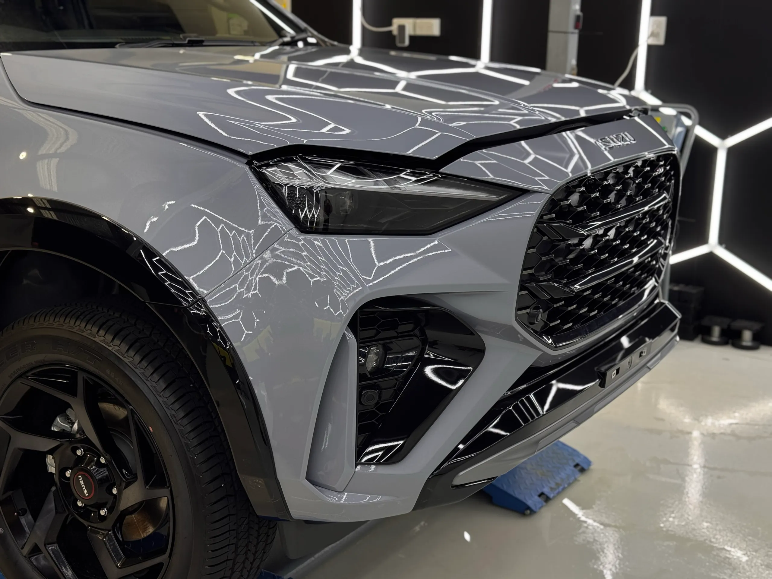 Grey Isuzu MU-X front wheel, headlight and grille beneath workshop lights during preparation