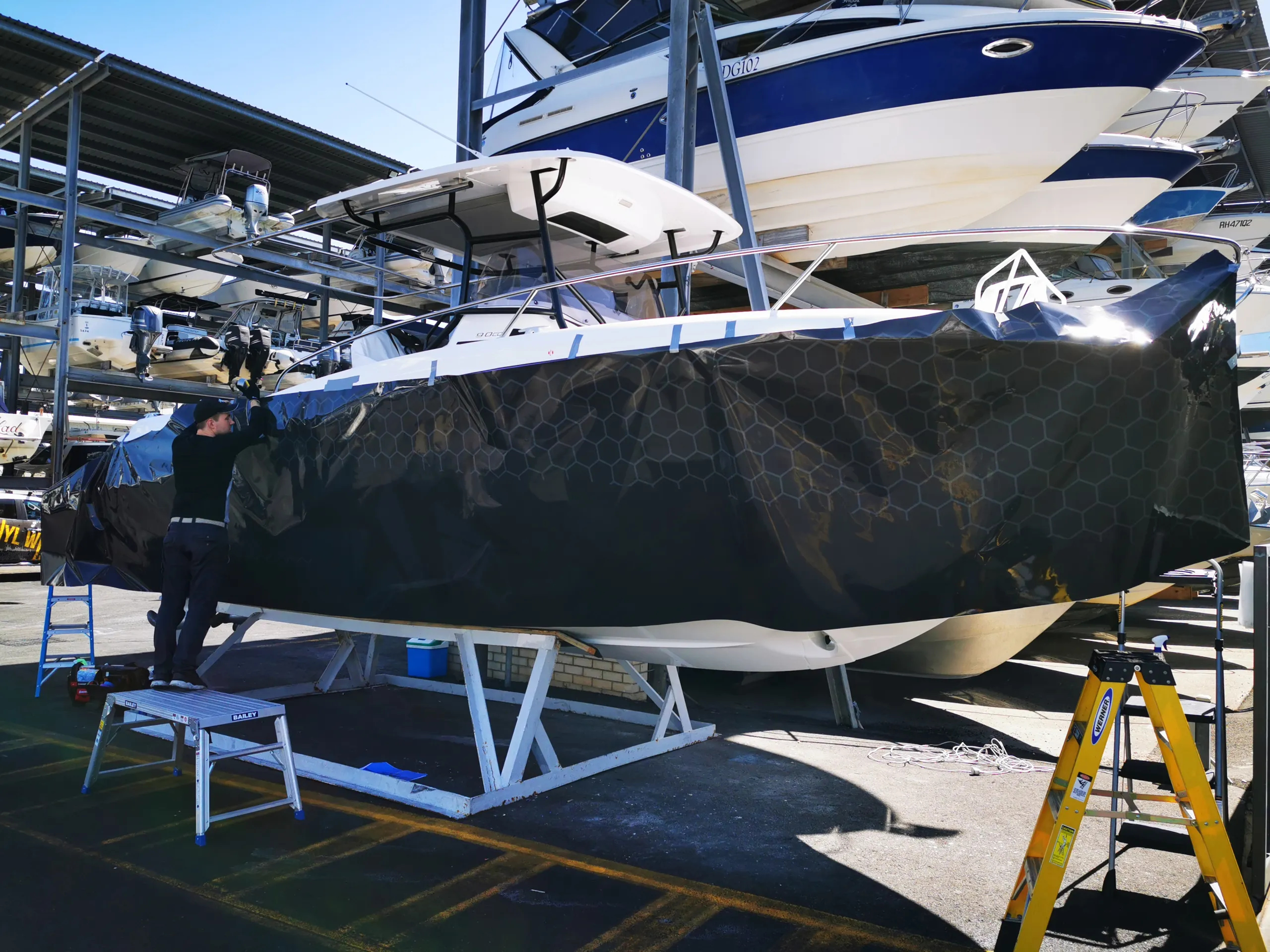 Installer applying dark honeycomb graphics along the Cap Camarat hull at the boat yard