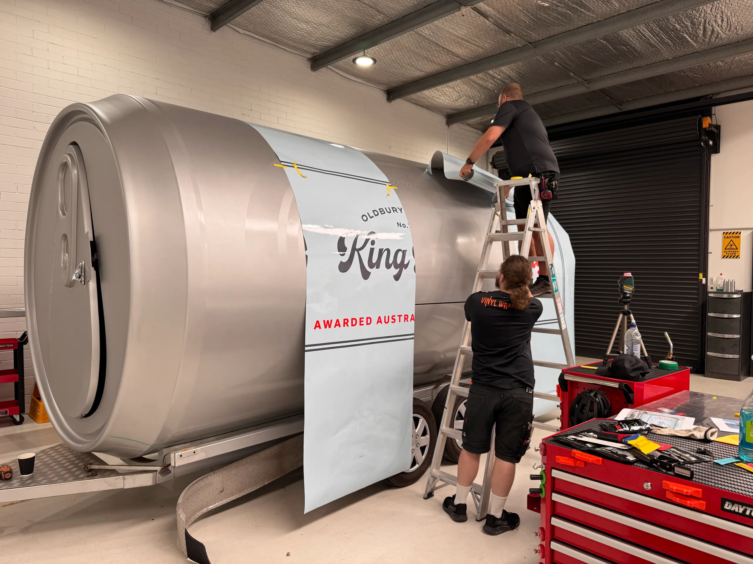 Back-facing installers positioning printed King Road Lager panels on the can-shaped trailer