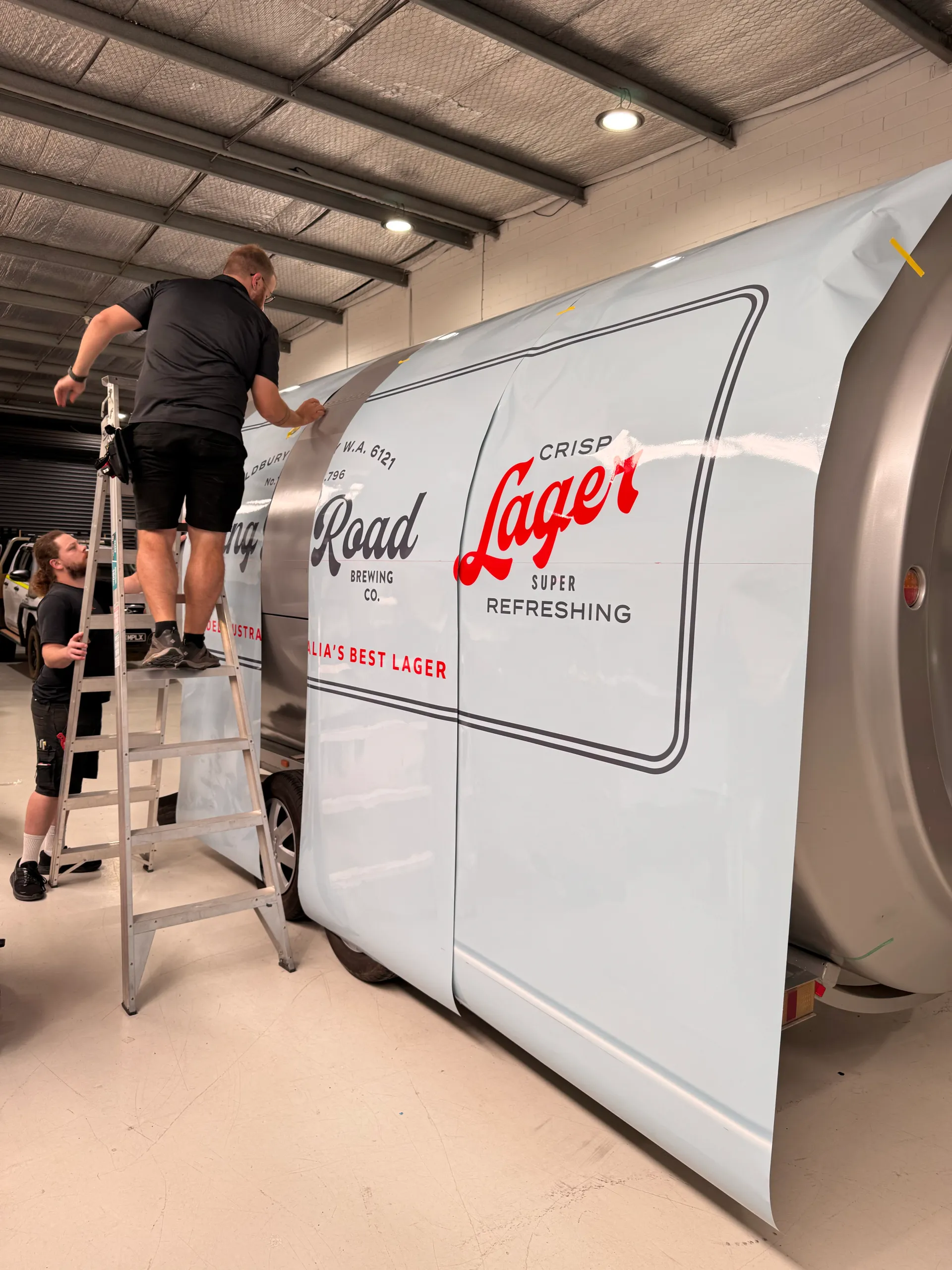 Installers applying King Road Lager graphics to the curved trailer side