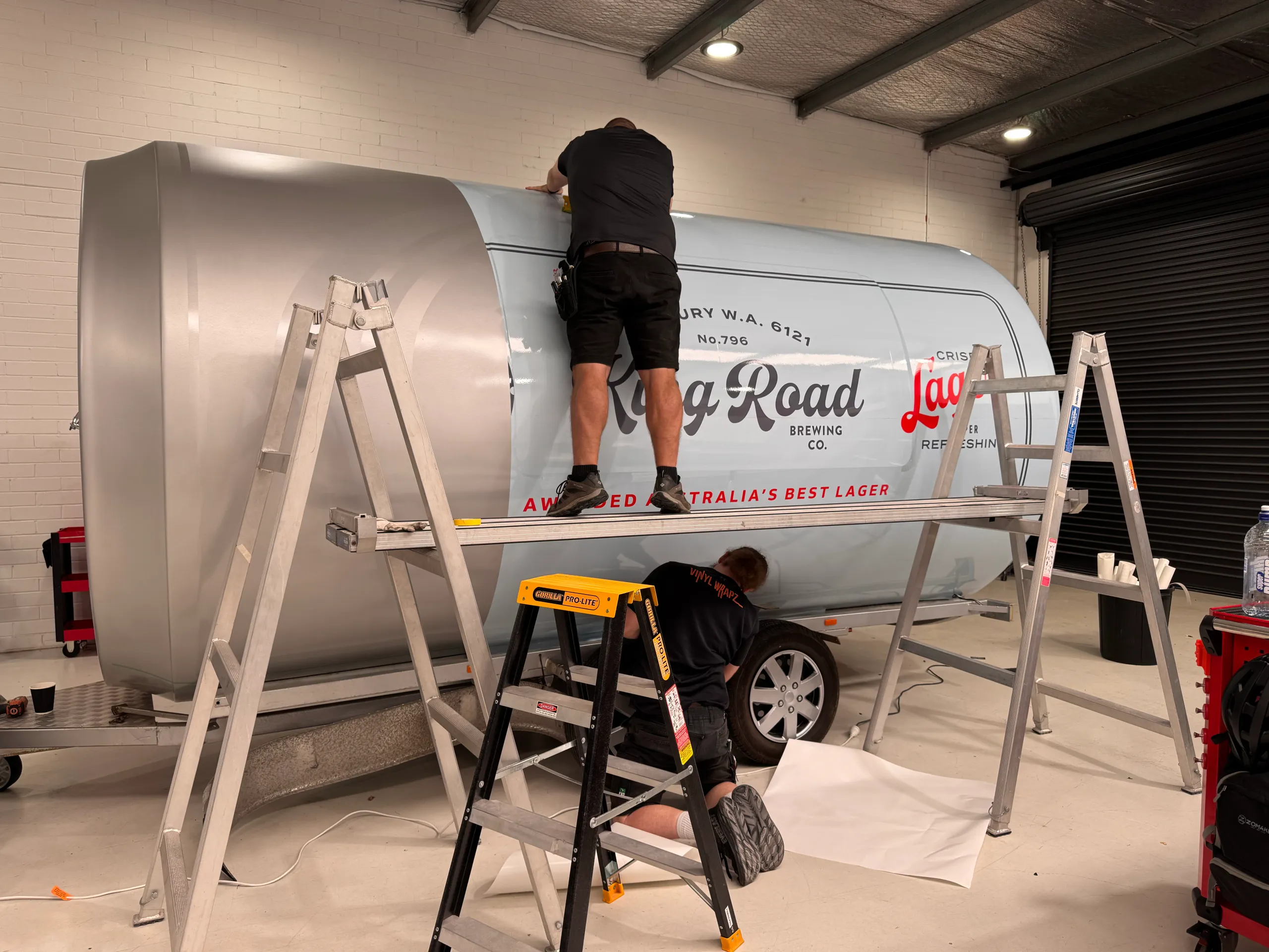 Back-facing installers fitting King Road Lager graphics around the can-shaped trailer