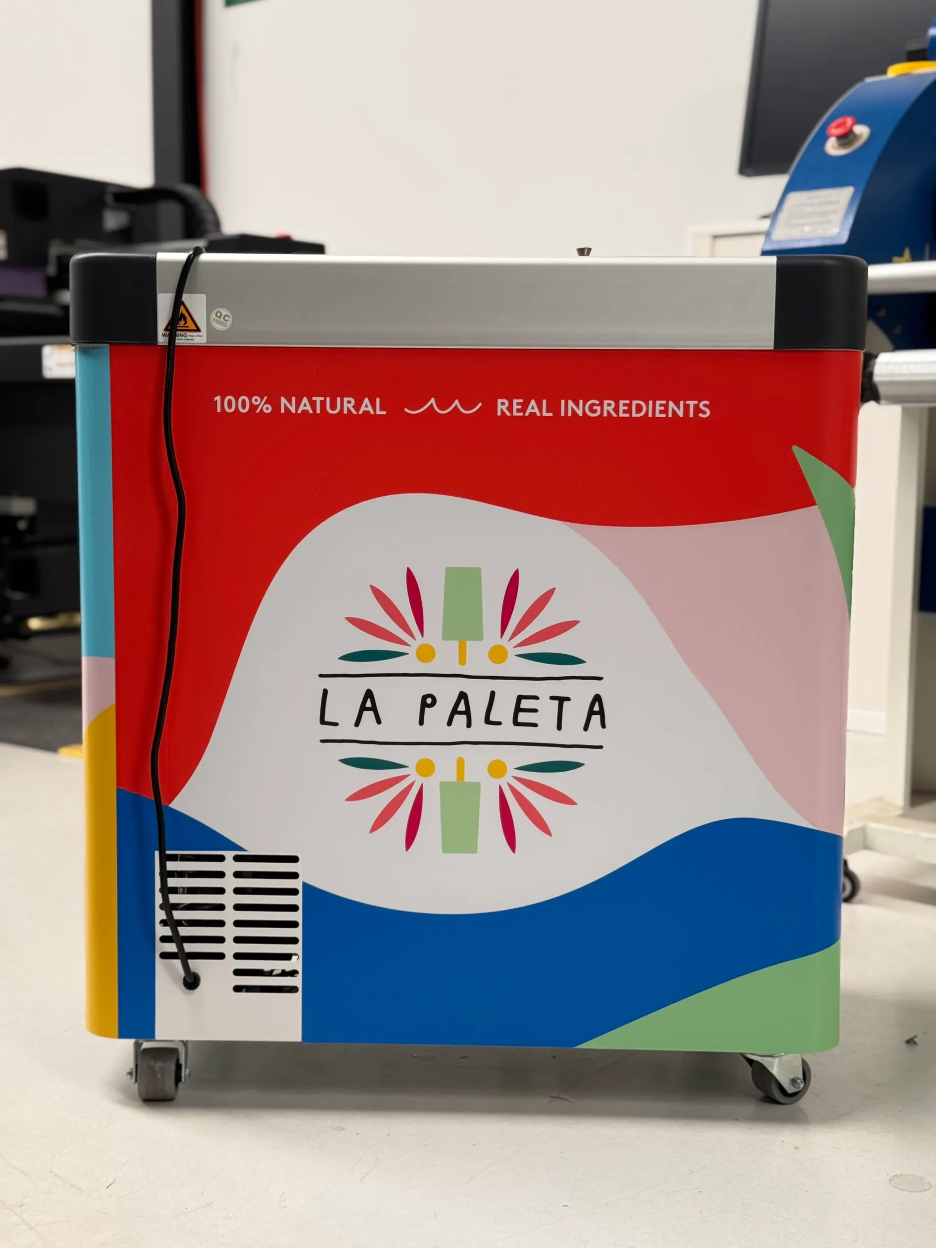 Colourful La Paleta graphics on the freezer rear panel beside its power cable and vent