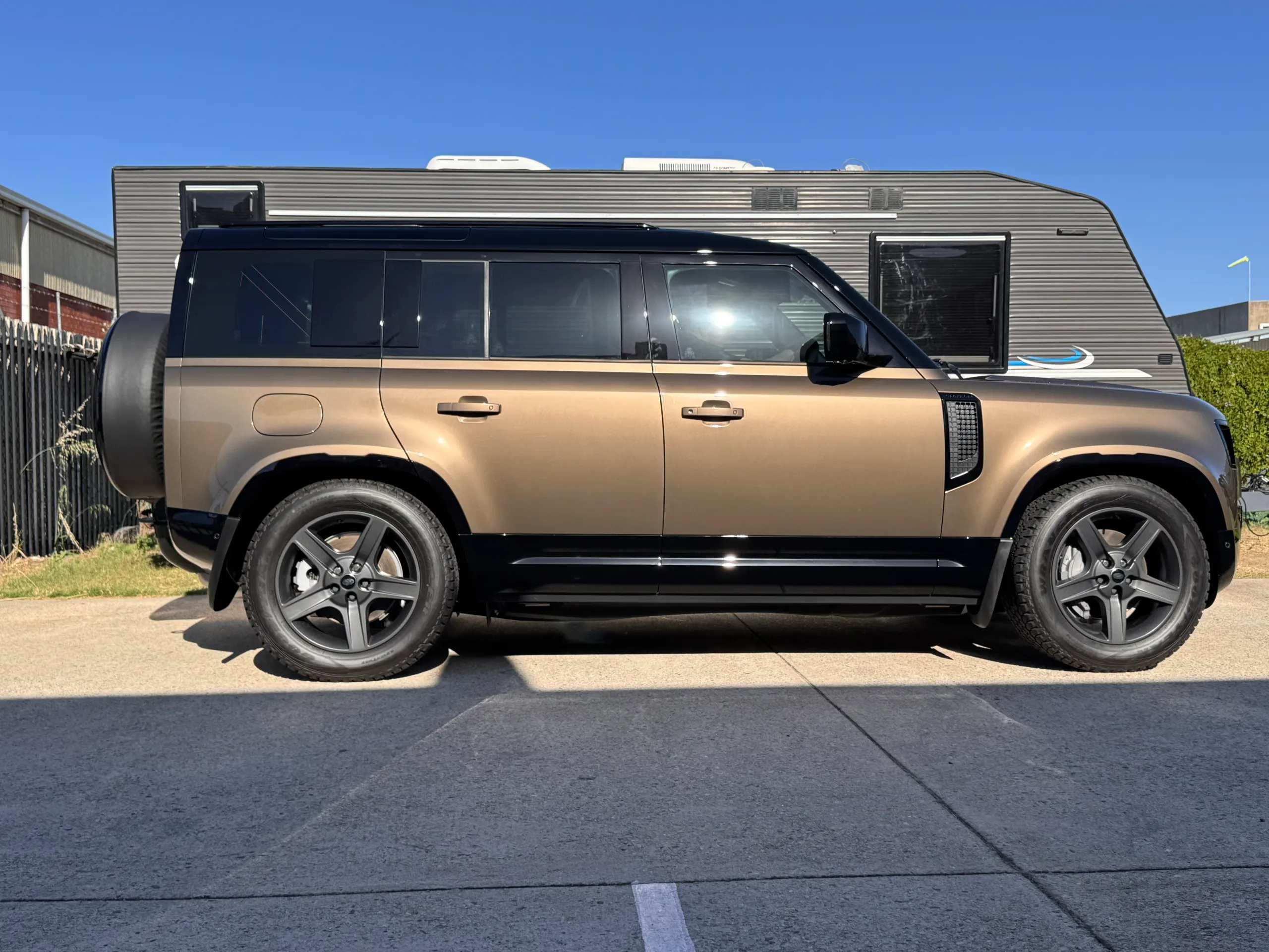 Side view of a bronze Land Rover Defender parked outdoors beside a caravan