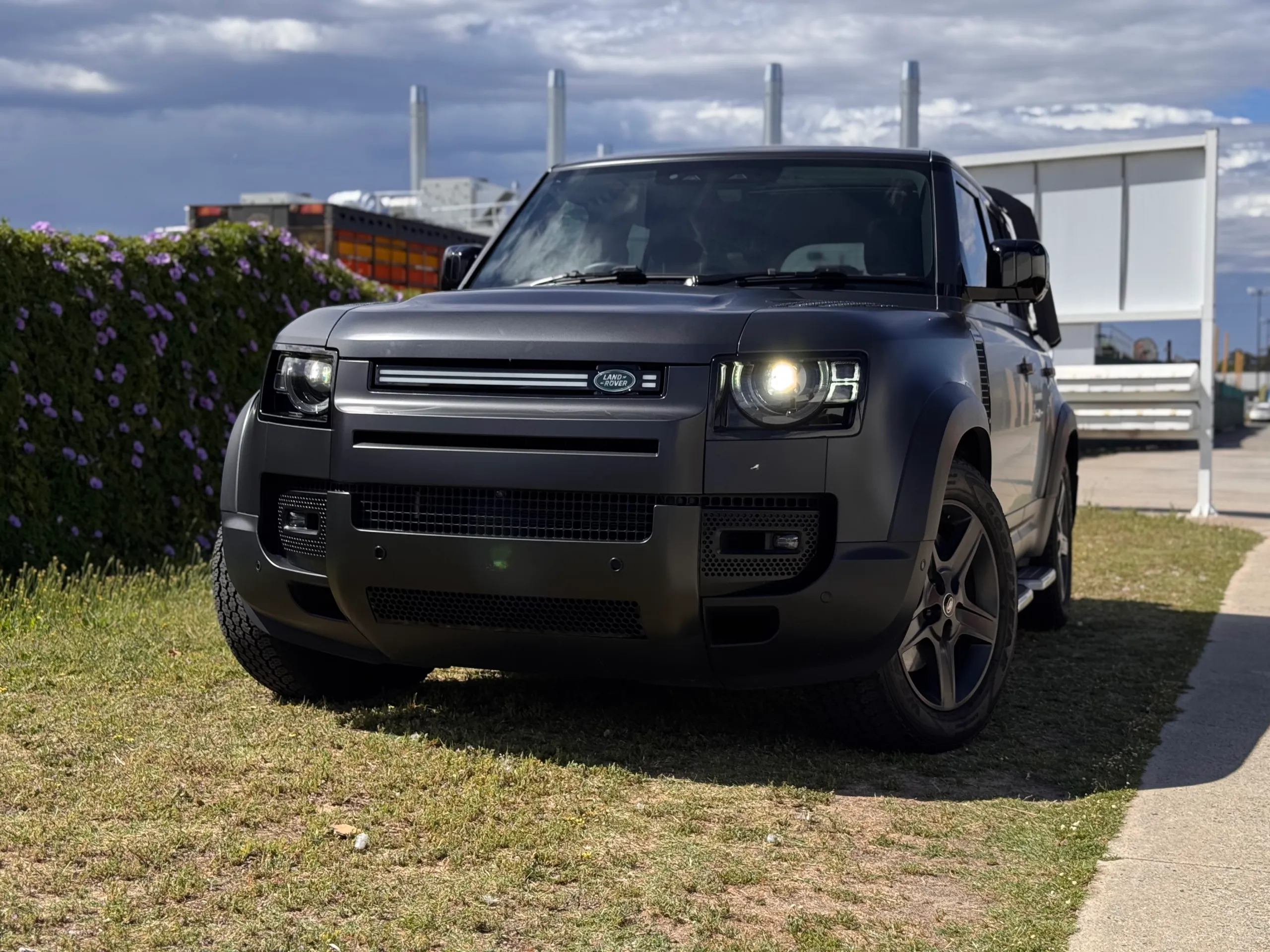 Front view of a matte dark grey Land Rover Defender parked on grass with its lights on
