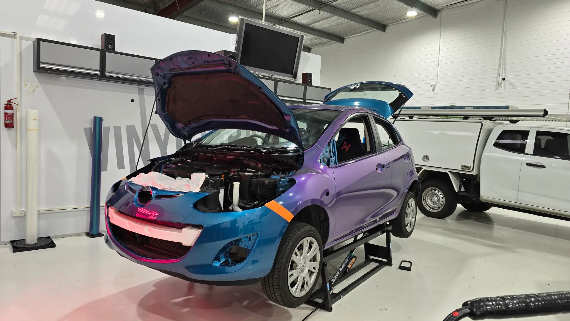 Turquoise and lavender Mazda 2 during wrapping in the workshop, front quarter