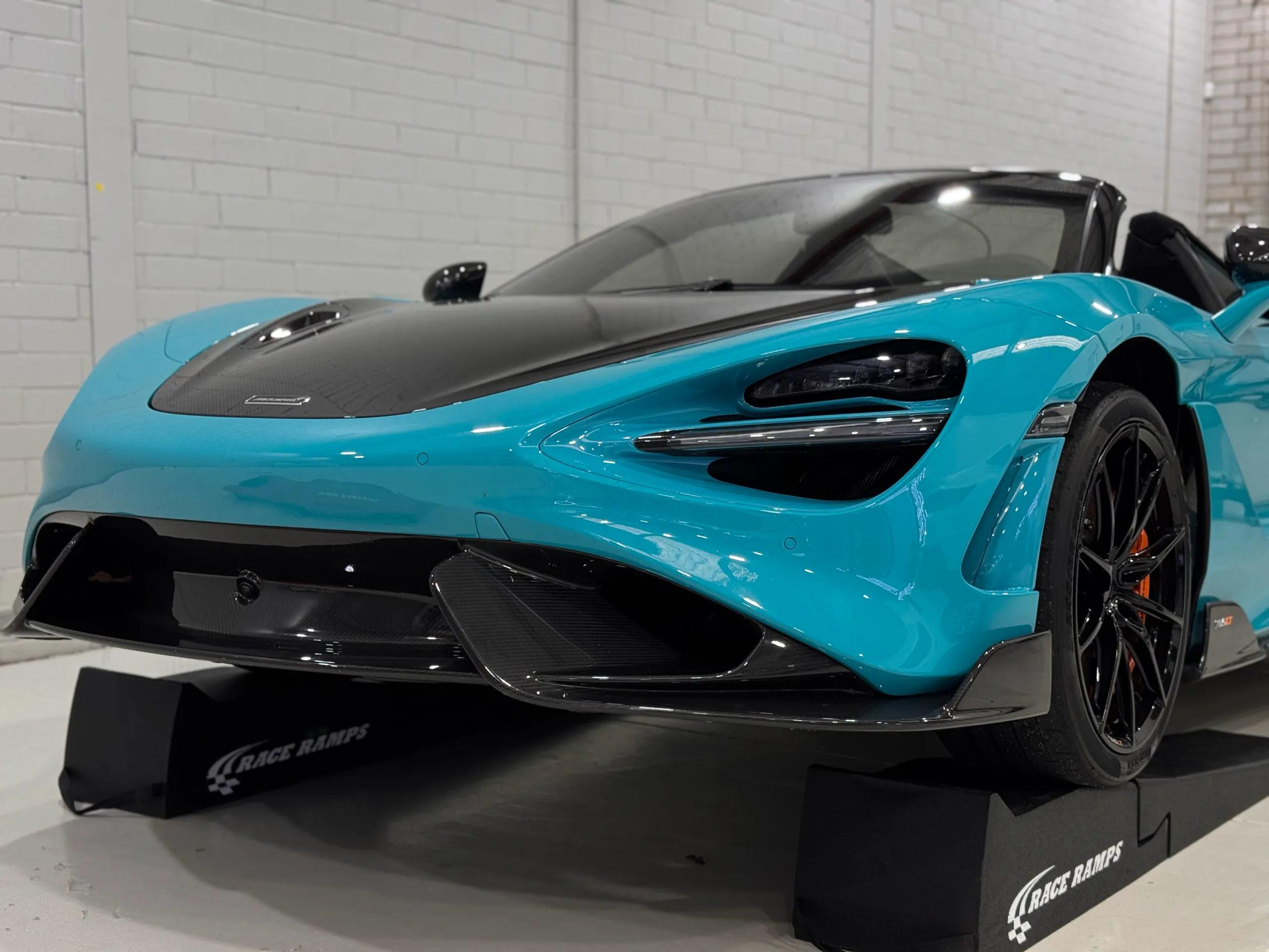 Low front view of a blue McLaren with a carbon bonnet on black workshop ramps