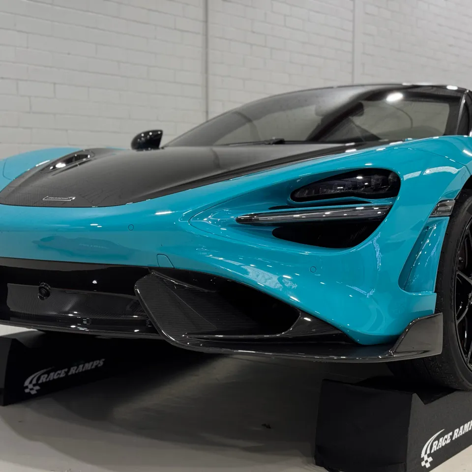 Low front view of a blue McLaren with a carbon bonnet on black workshop ramps