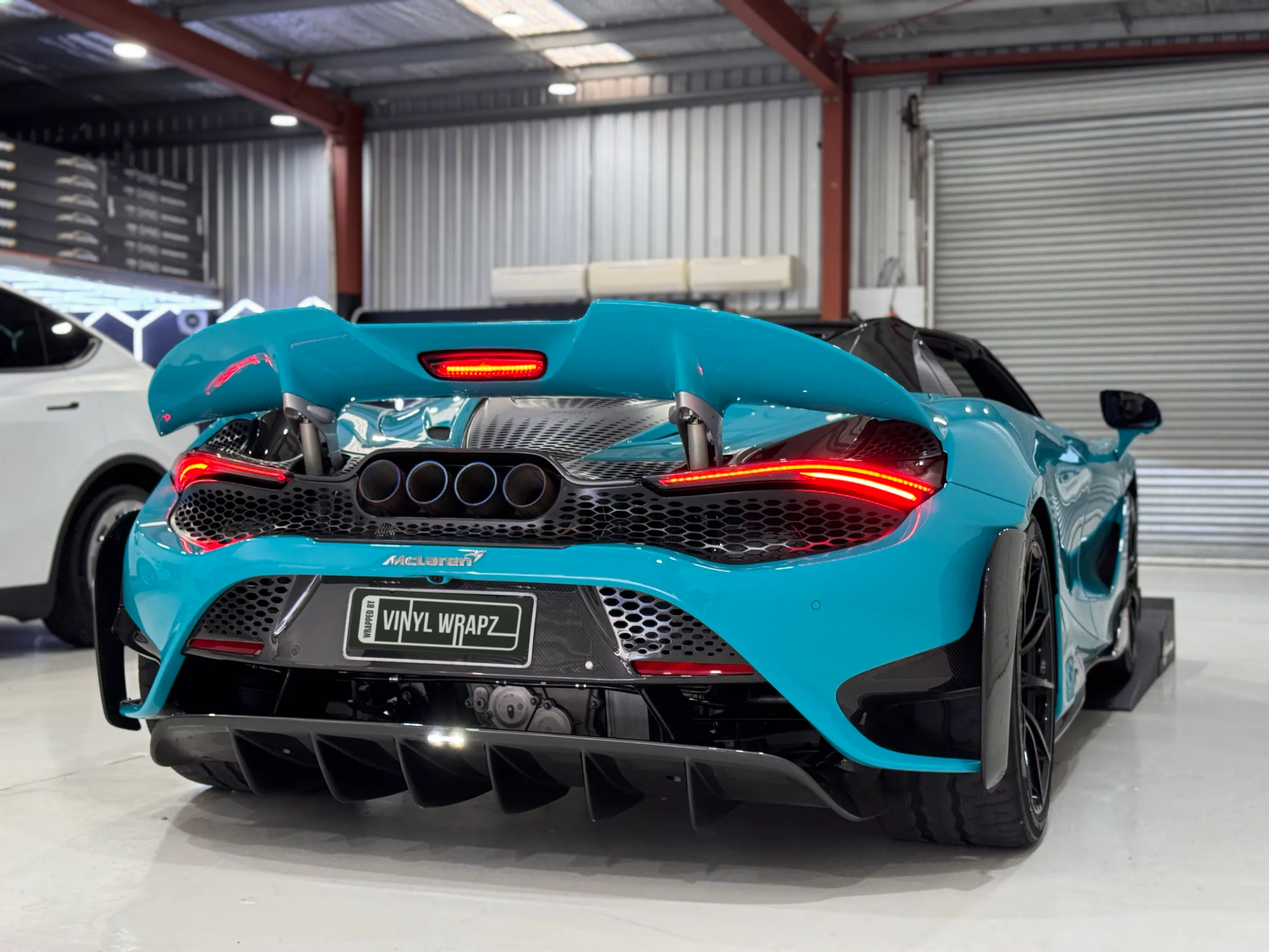 Rear view of a blue McLaren with its wing raised, triple exhaust outlets and illuminated tail lights