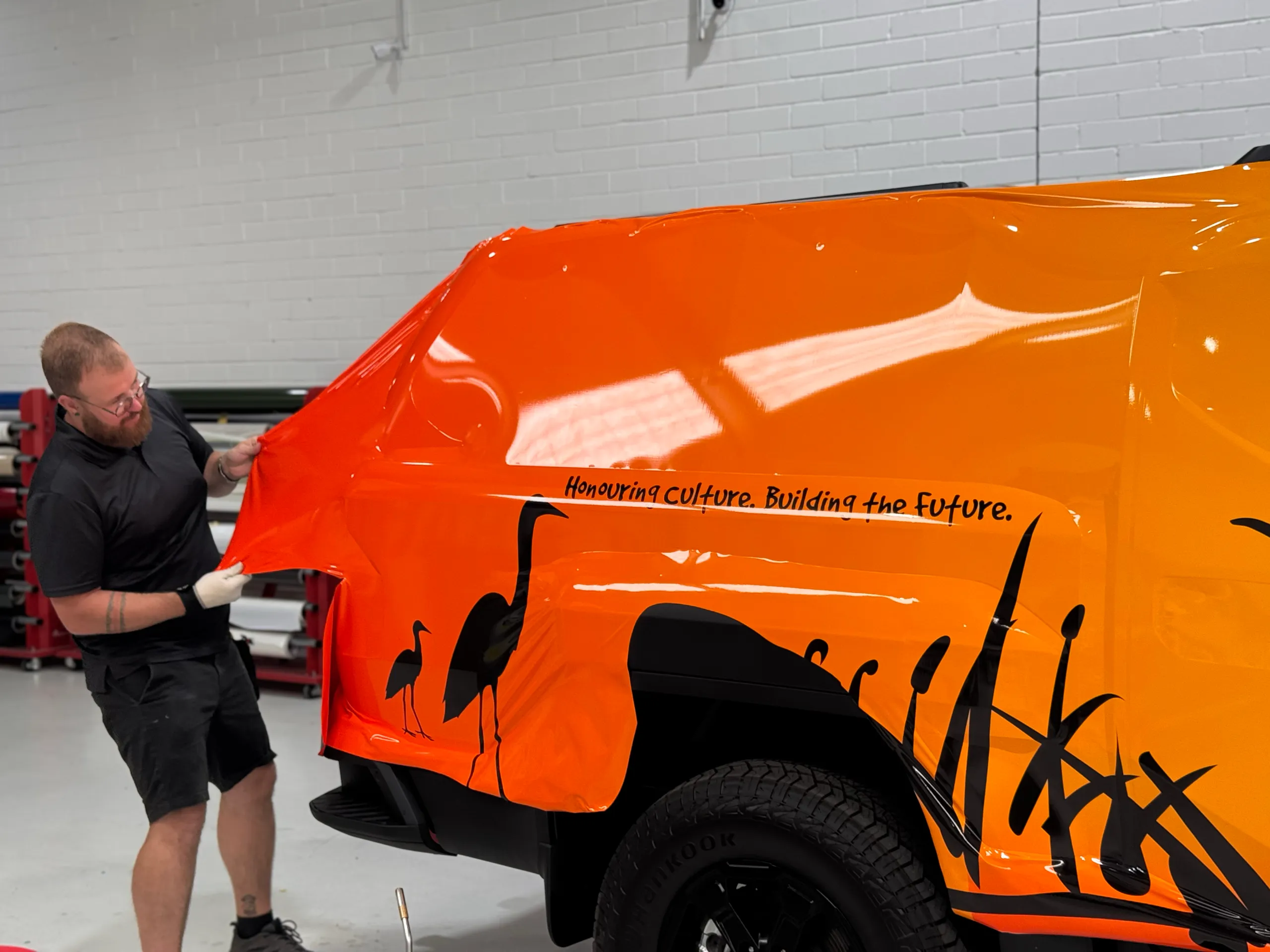 Installer fitting orange Milliyaan graphics with black bird silhouettes to a Kia Tasman rear quarter
