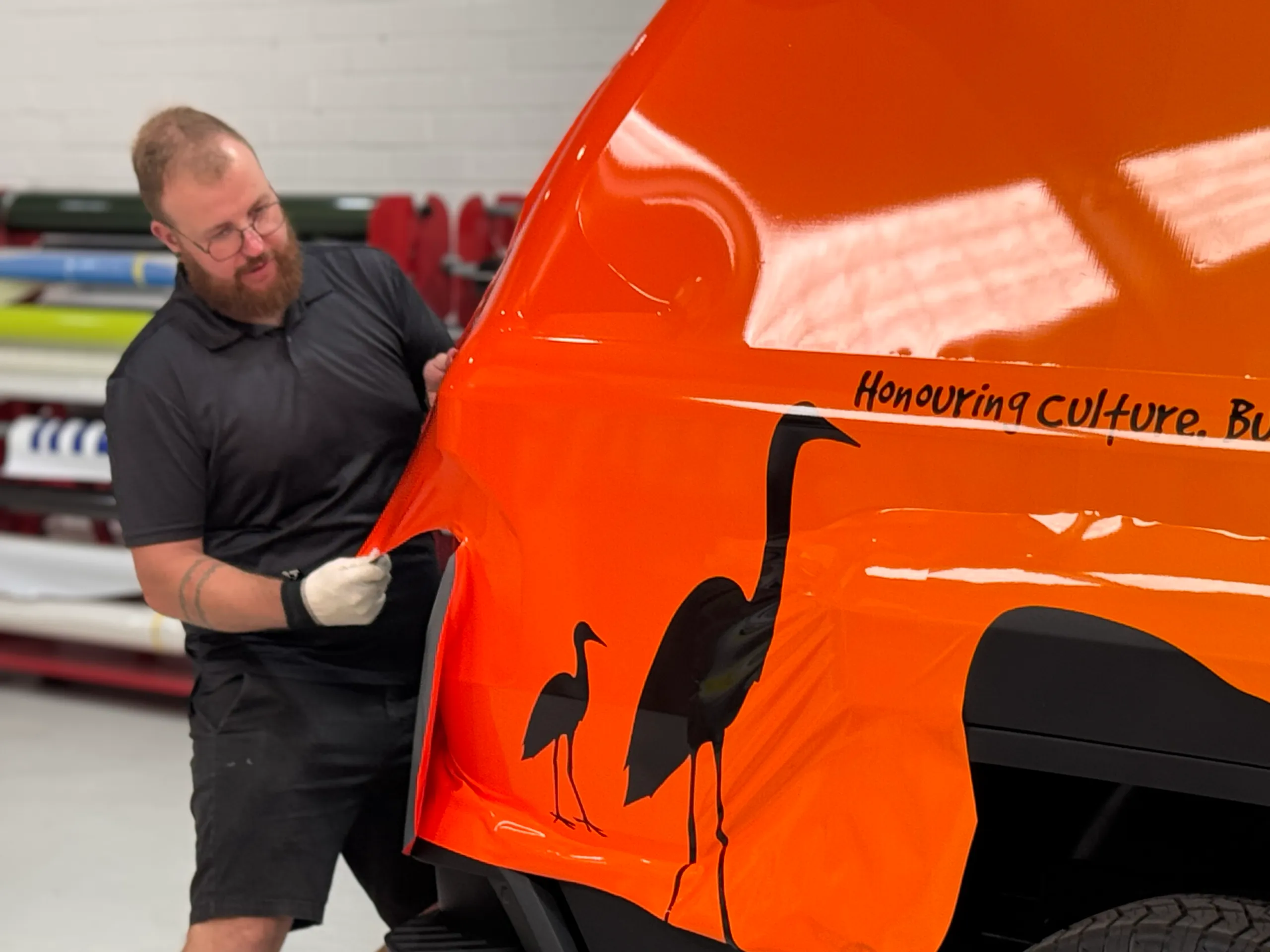 Orange printed wrap being shaped around the Kia Tasman rear quarter by an installer
